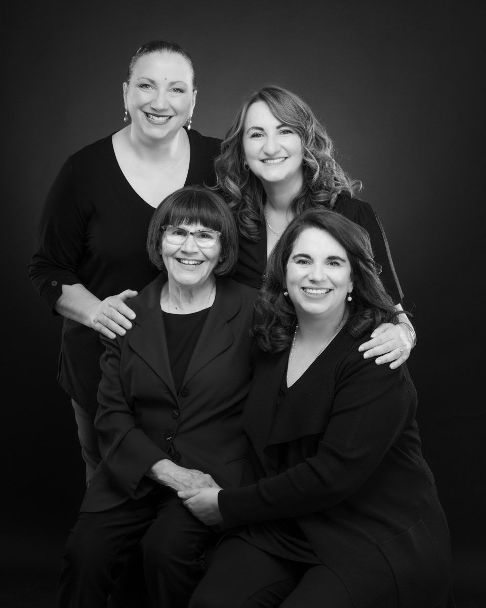 A professional black and white studio portrait of four women in dark attire smiling together against a dark background.