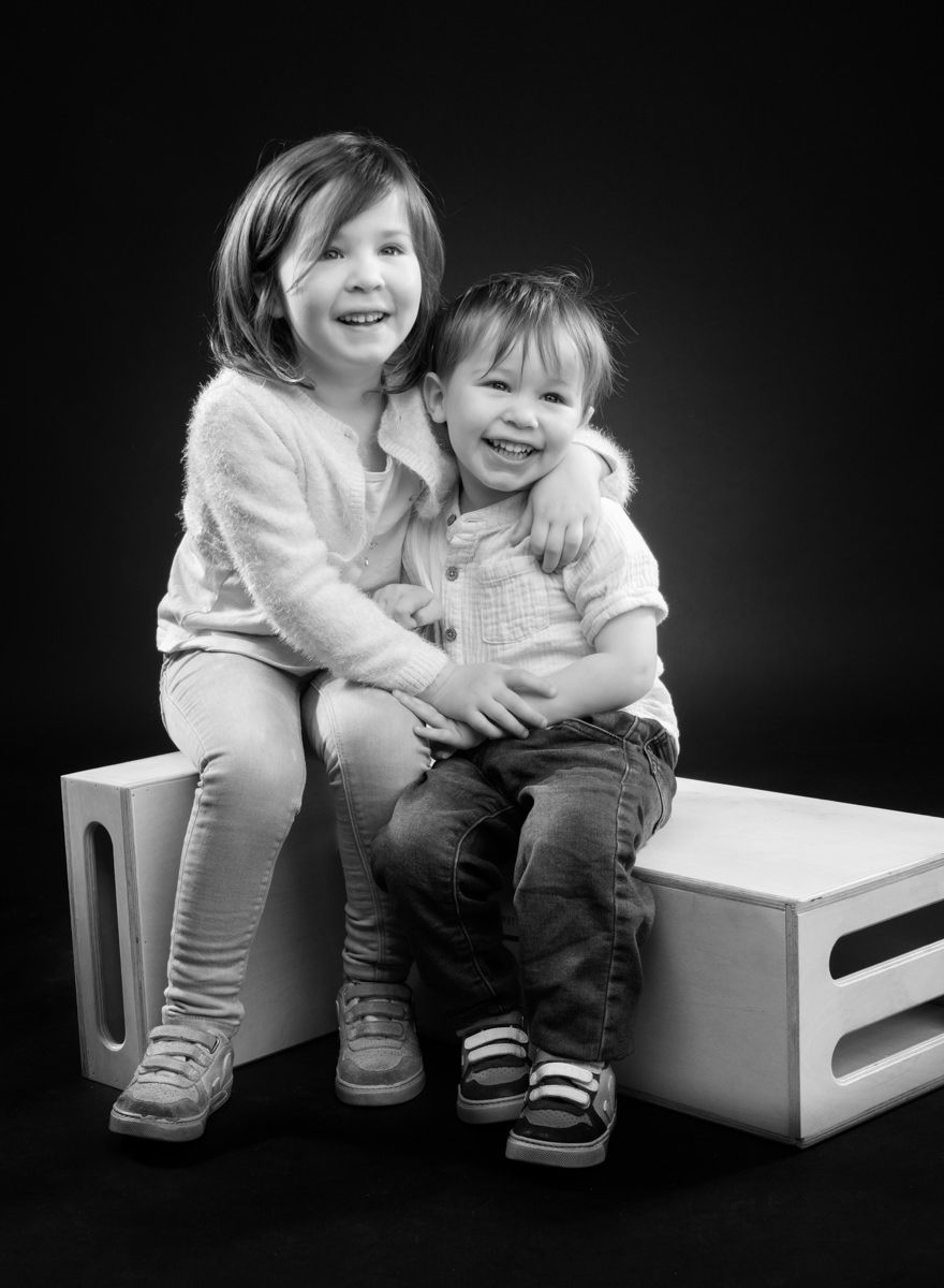 Black and white portrait series of two children laughing and hugging while sitting on a white bench.