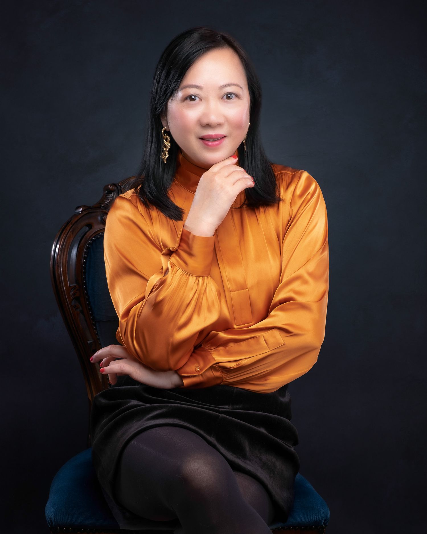 Professional portrait against dark background features person in orange silk blouse and black skirt seated in vintage chair.