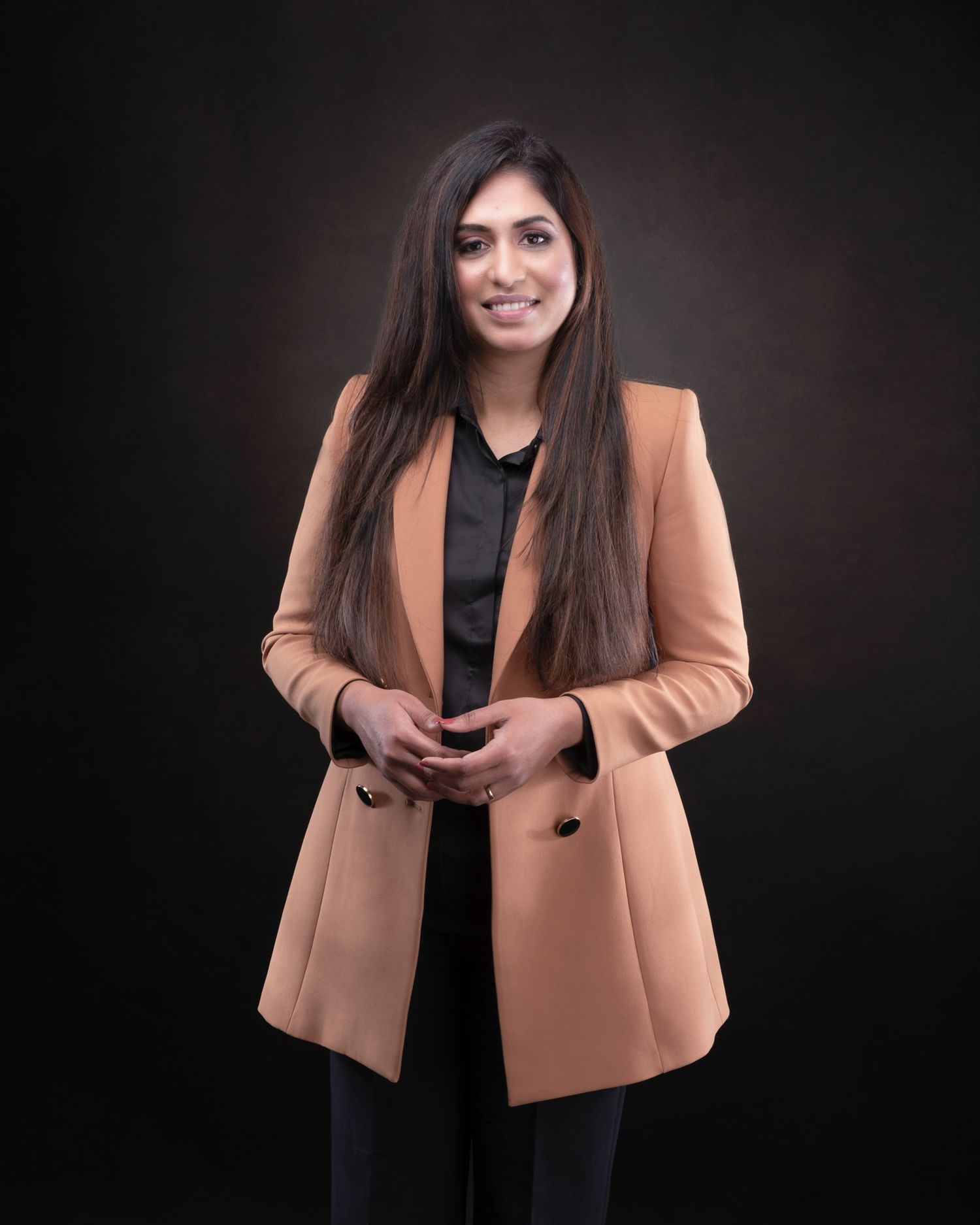 Professional portrait of person wearing a camel colored blazer over black attire against dark background.