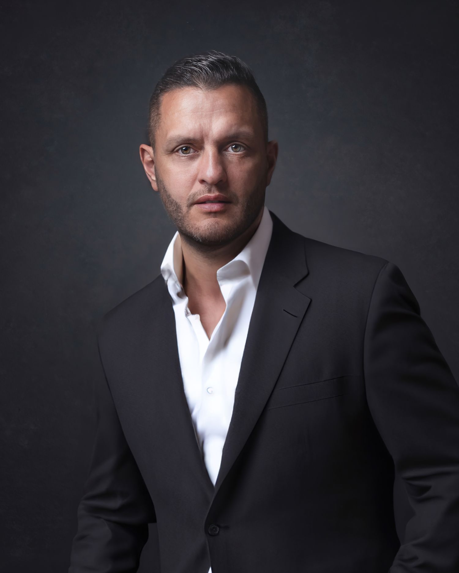 Professional studio portrait of a person in a dark suit jacket and white dress shirt against a dramatic dark background.