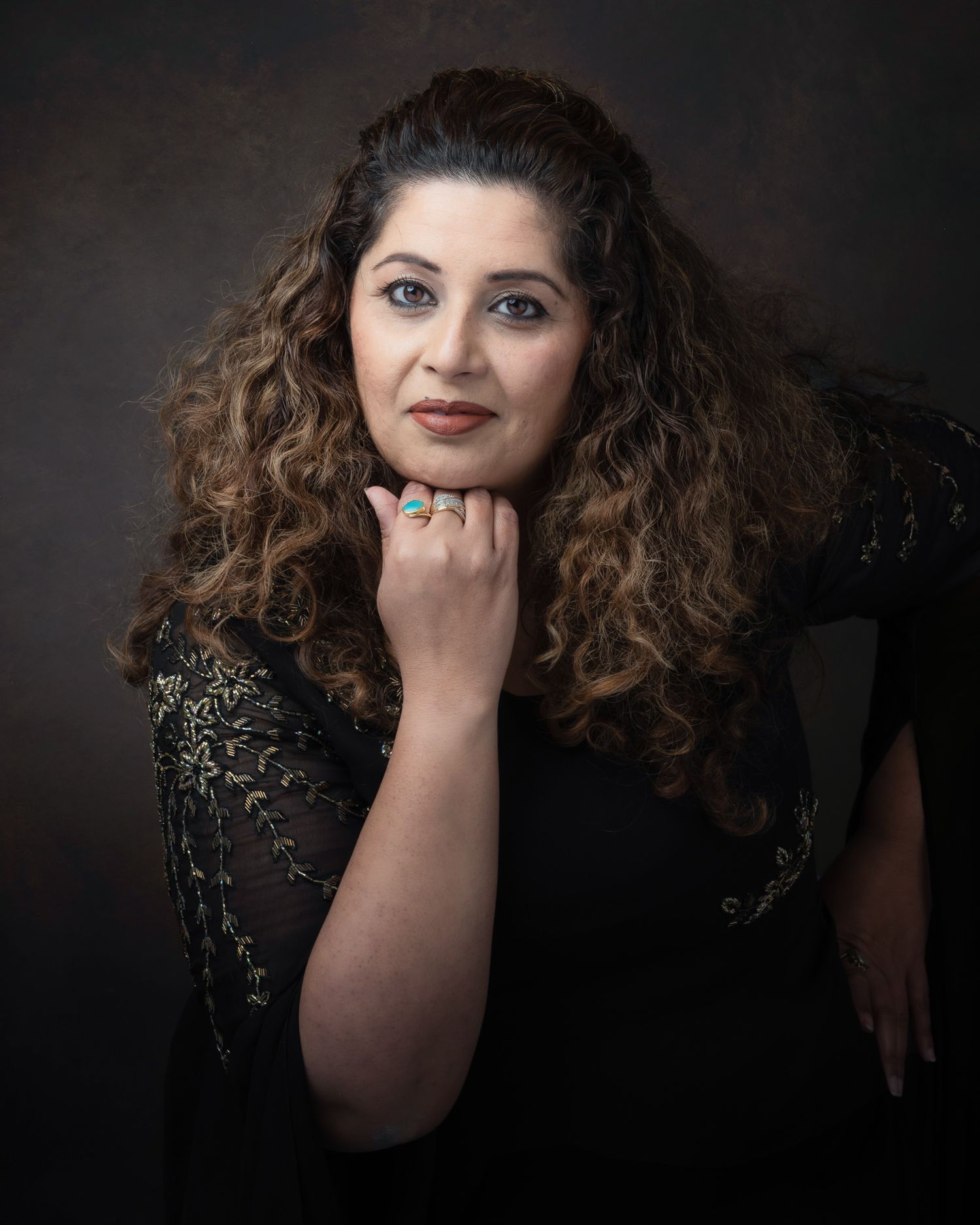 Studio portrait of a person with long curly dark hair in black lace against dark background resting hand under chin.