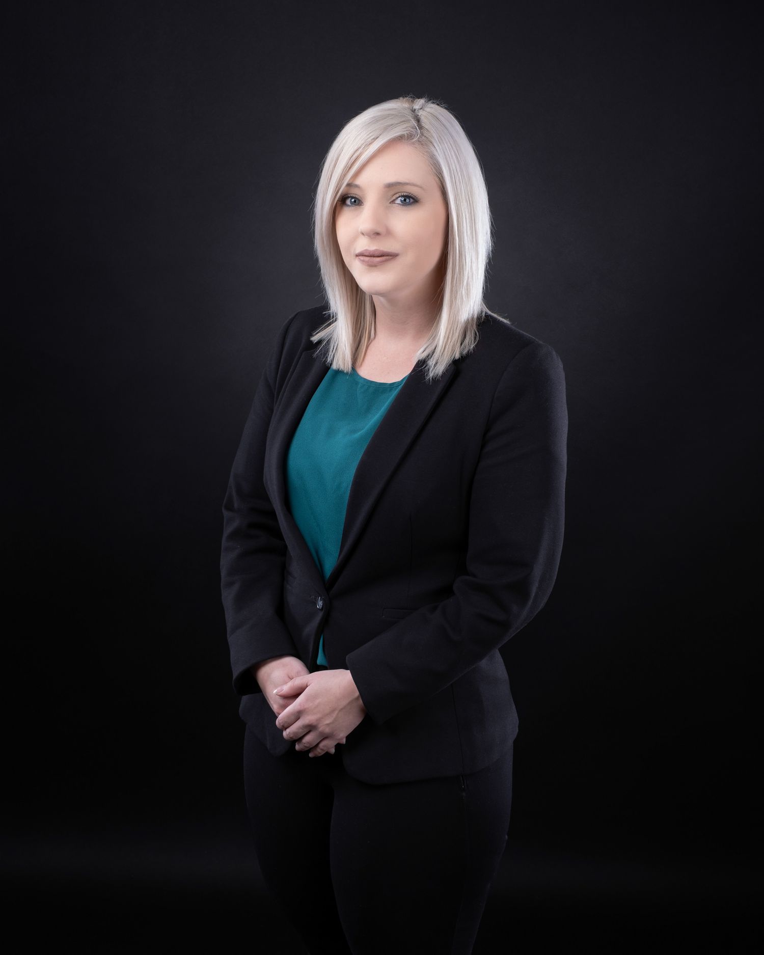 Professional studio portrait of a person in a black blazer and teal blouse against a dark background.
