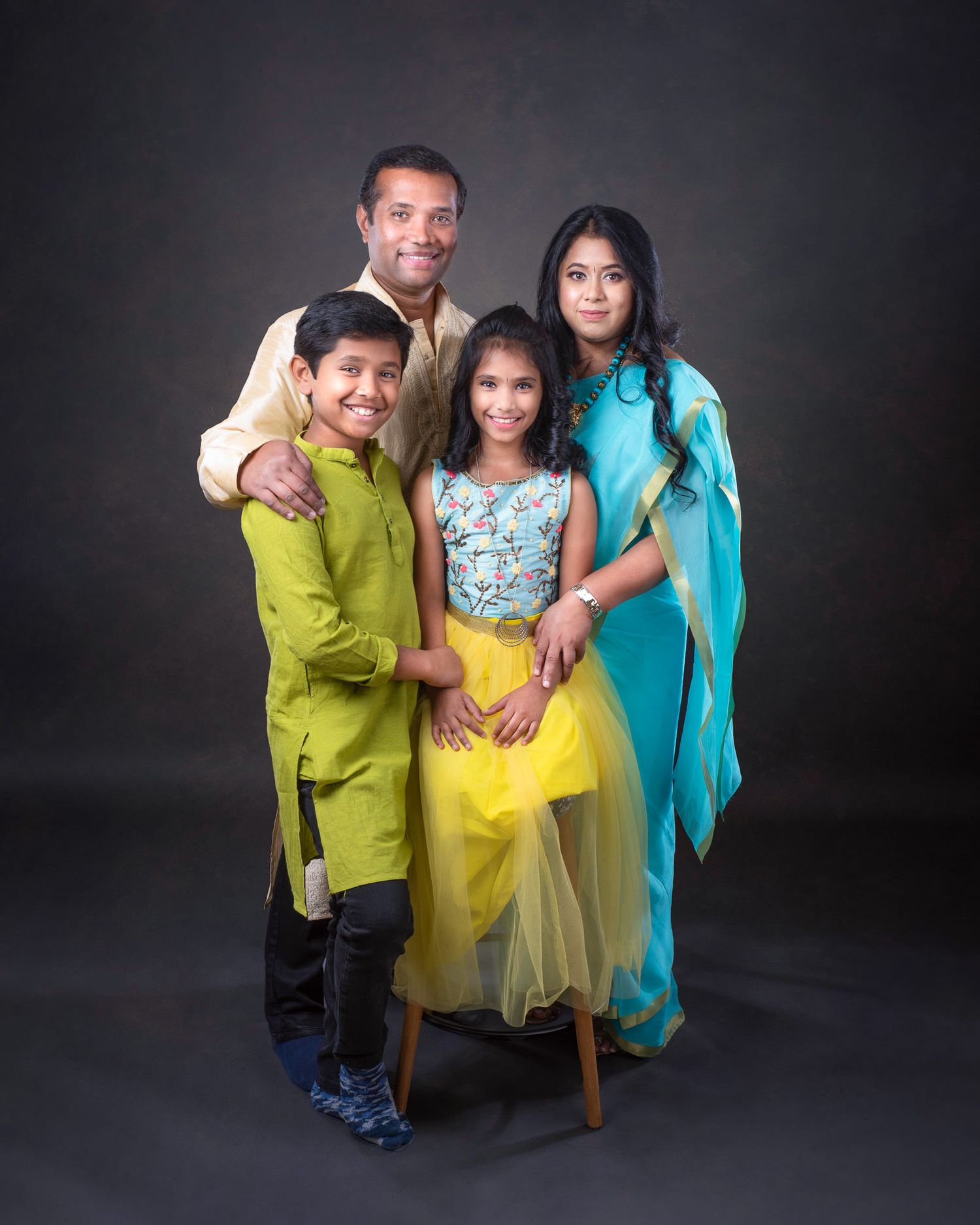 A family of four wearing vibrant traditional Indian-style clothing poses together against a dark backdrop in a studio portrait.