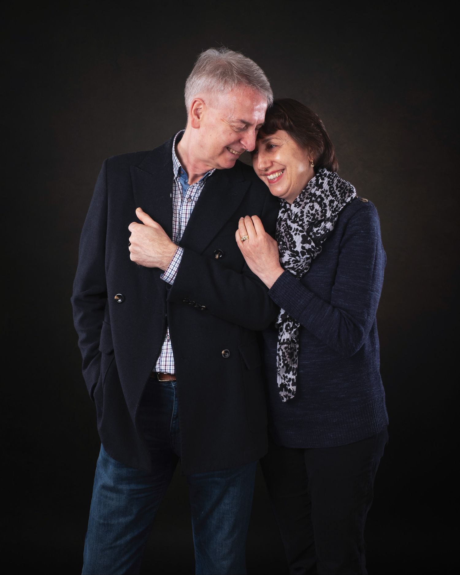 A couple embraces while sharing a tender moment together against a dark background, dressed in winter coats and scarves.