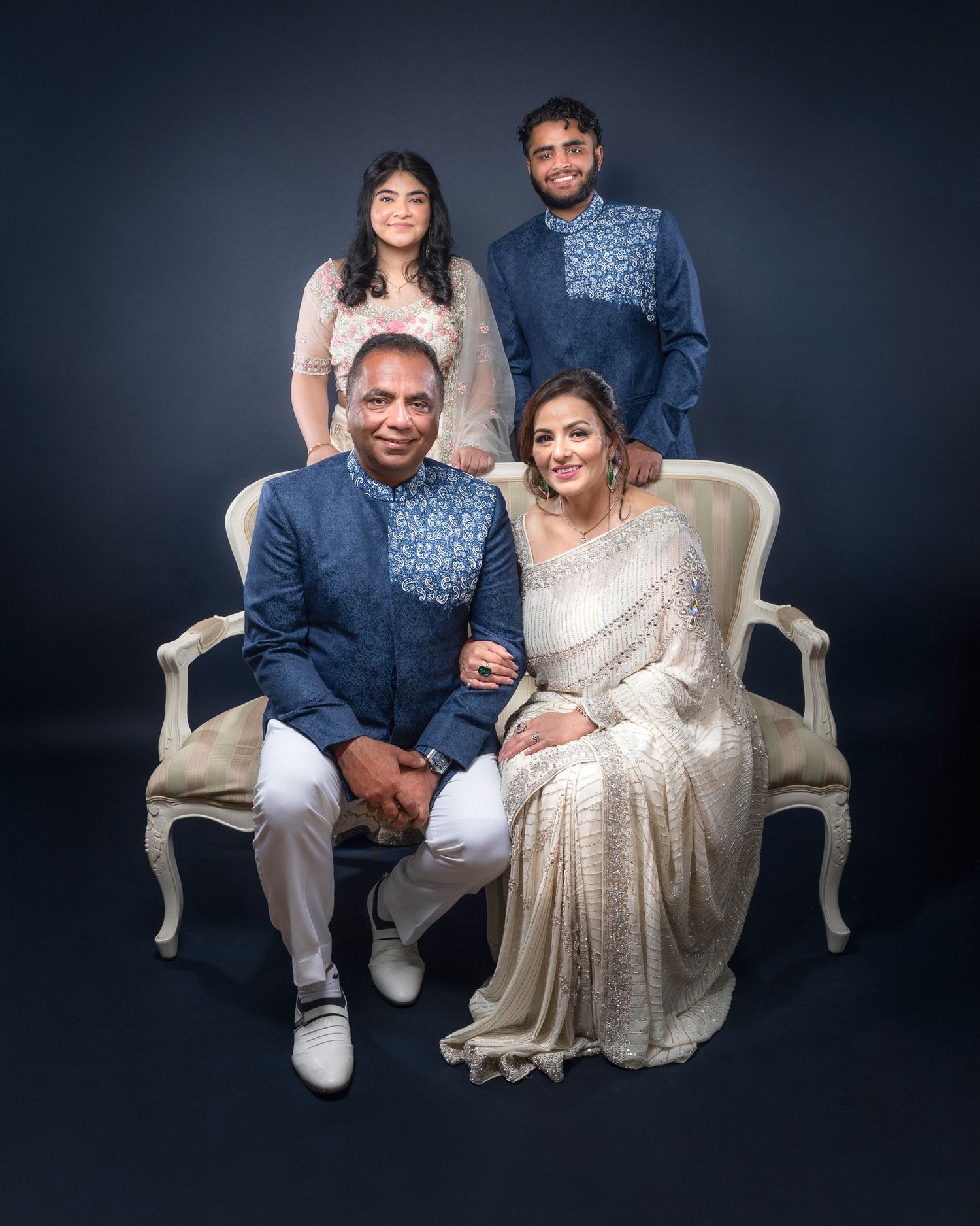 A formal family portrait with coordinated blue and white traditional Indian attire seated on an elegant cream settee.