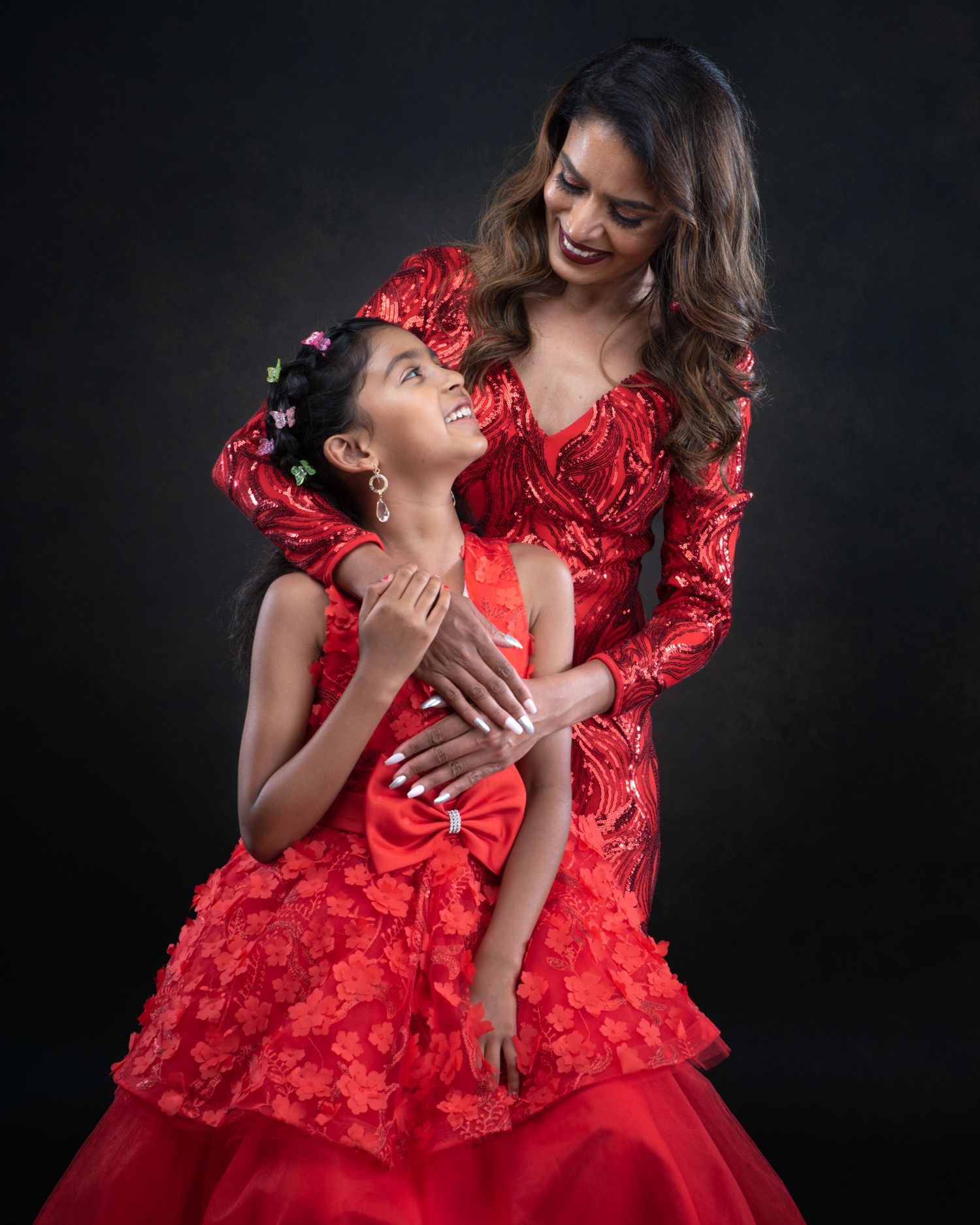 Two figures in flowing red gowns pose together dramatically against a dark background for a glamorous portrait.