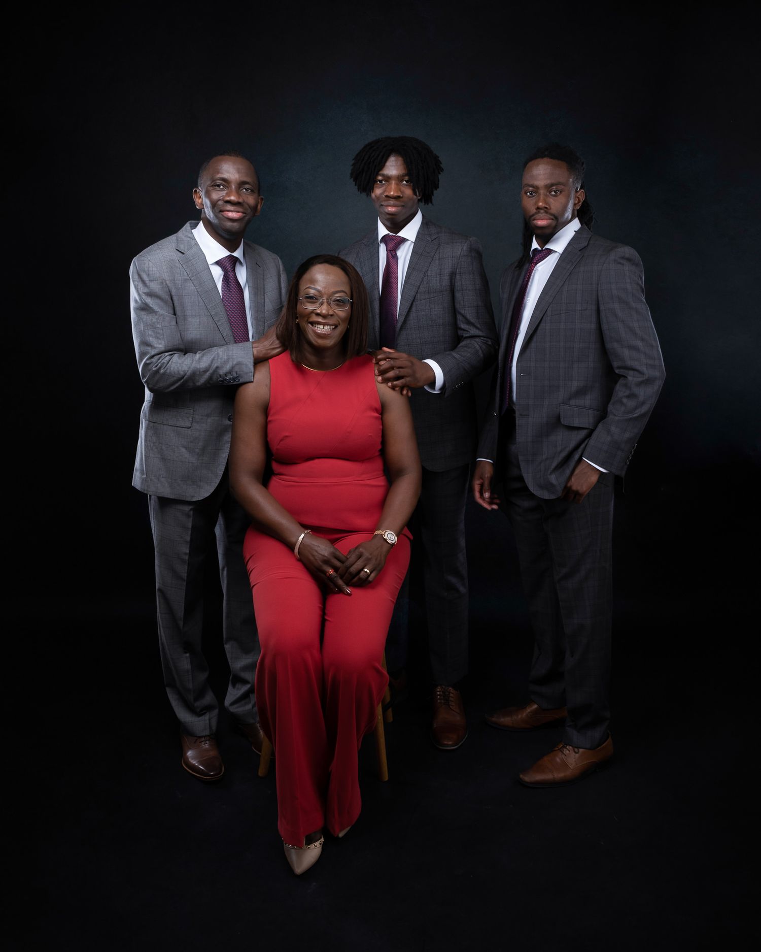 Professional studio portrait of a group in formal attire including a woman in red and three men in suits against dark background.