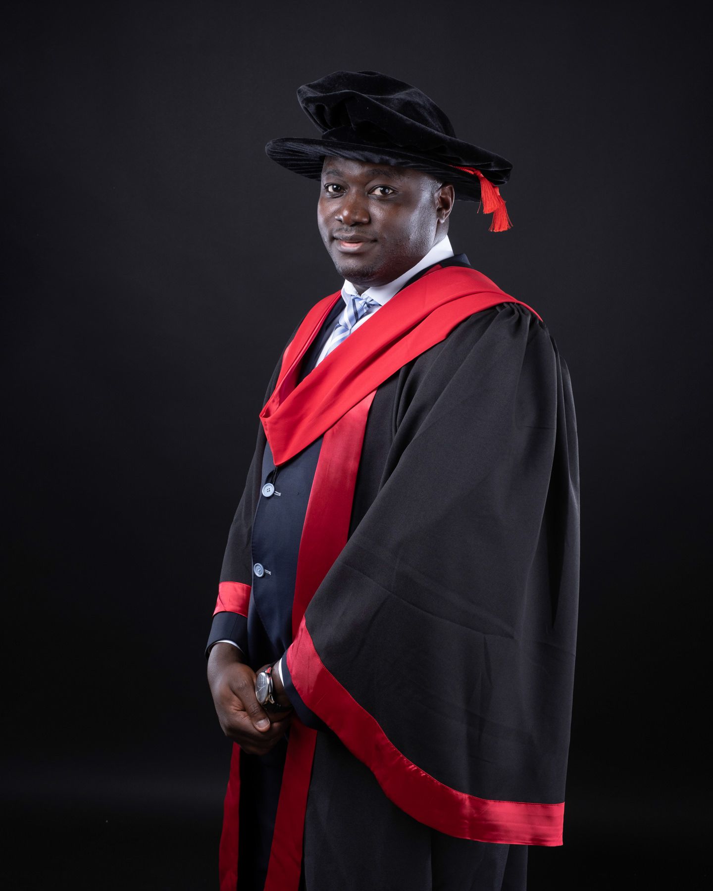 Graduate in traditional black academic gown and red stole poses against dark background in professional portrait series.