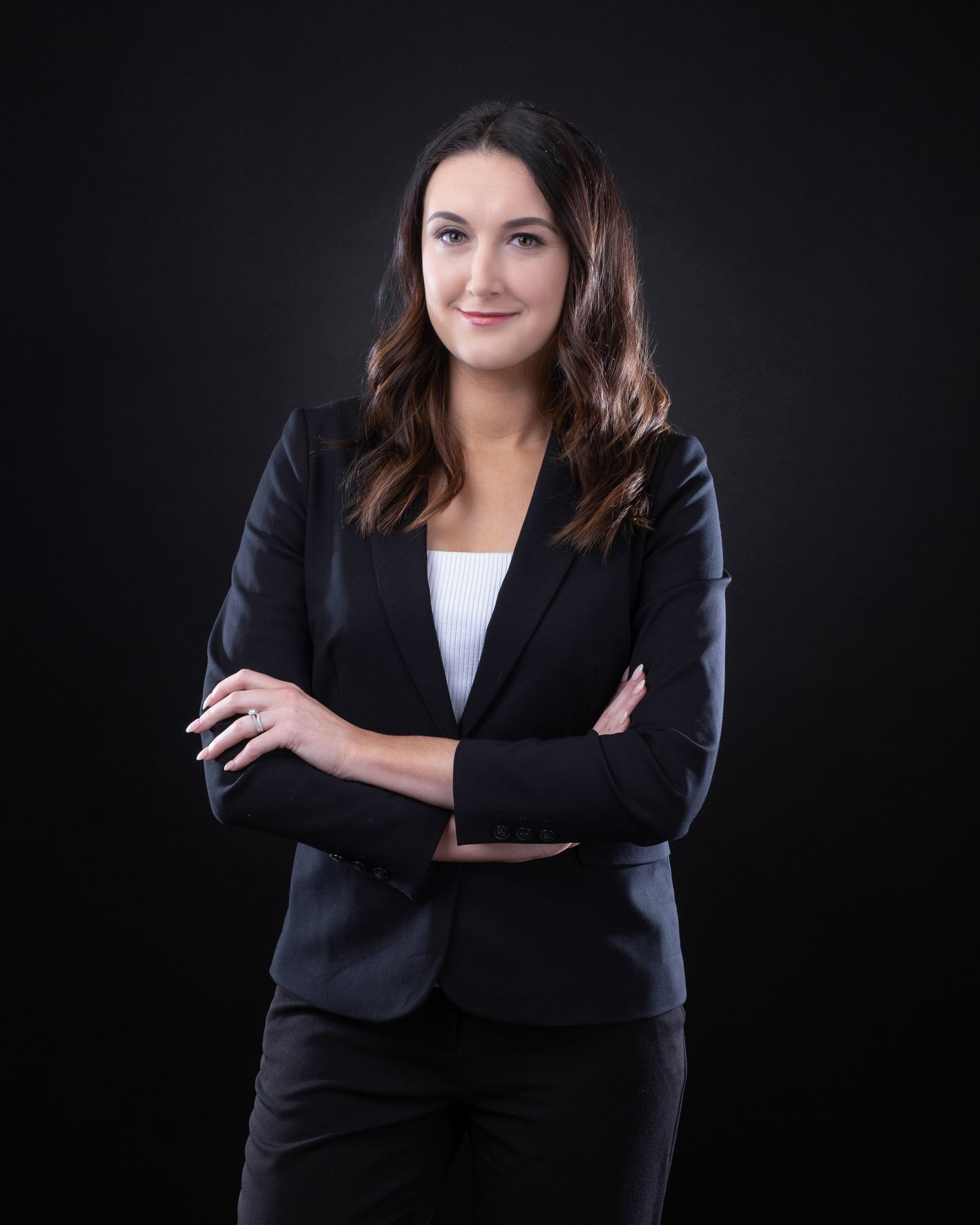 Professional business portrait of someone in a black blazer and white top against a dark background, arms crossed confidently.