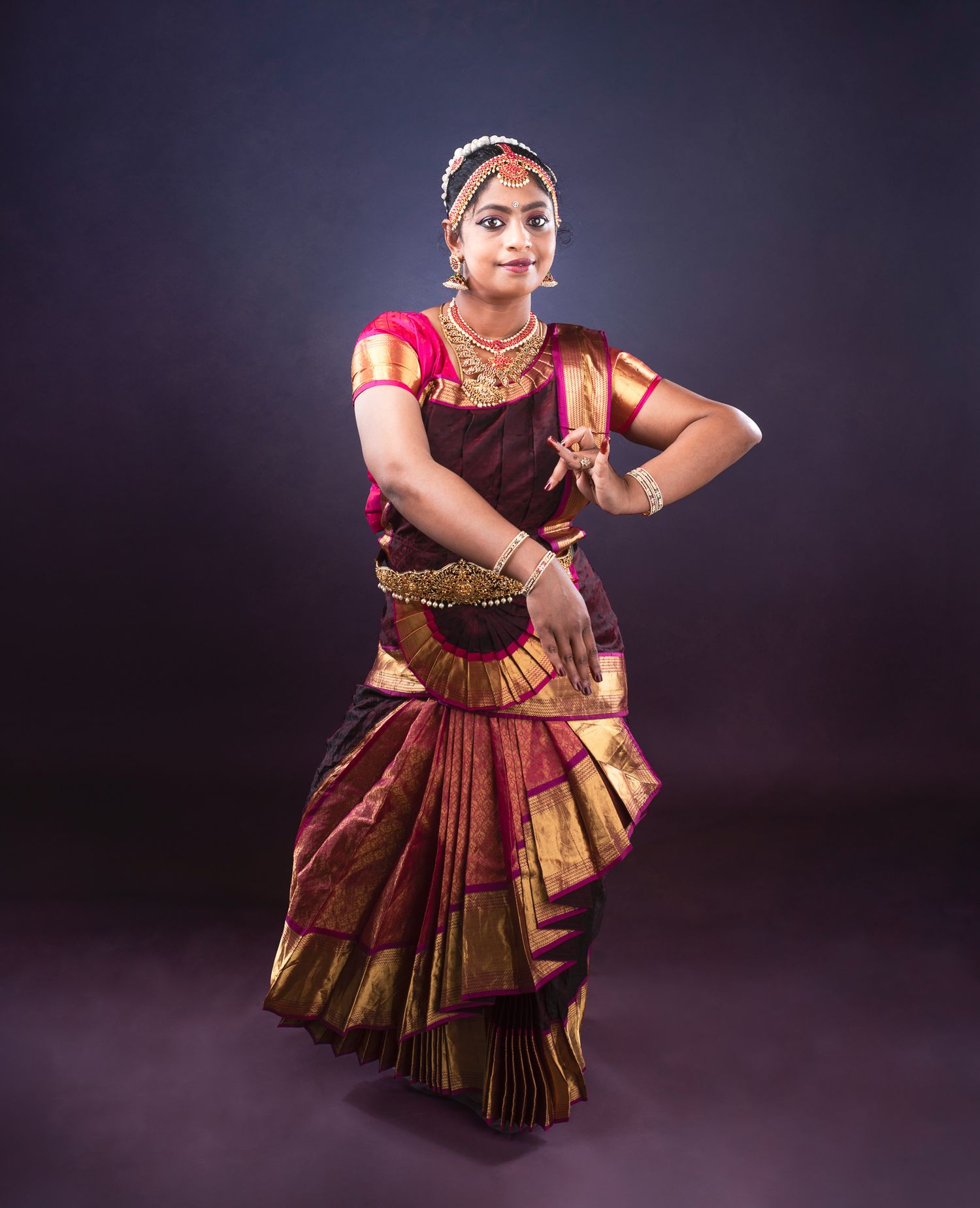 Classical Indian dancer in traditional red and gold Bharatanatyam costume performs graceful pose against dark backdrop.