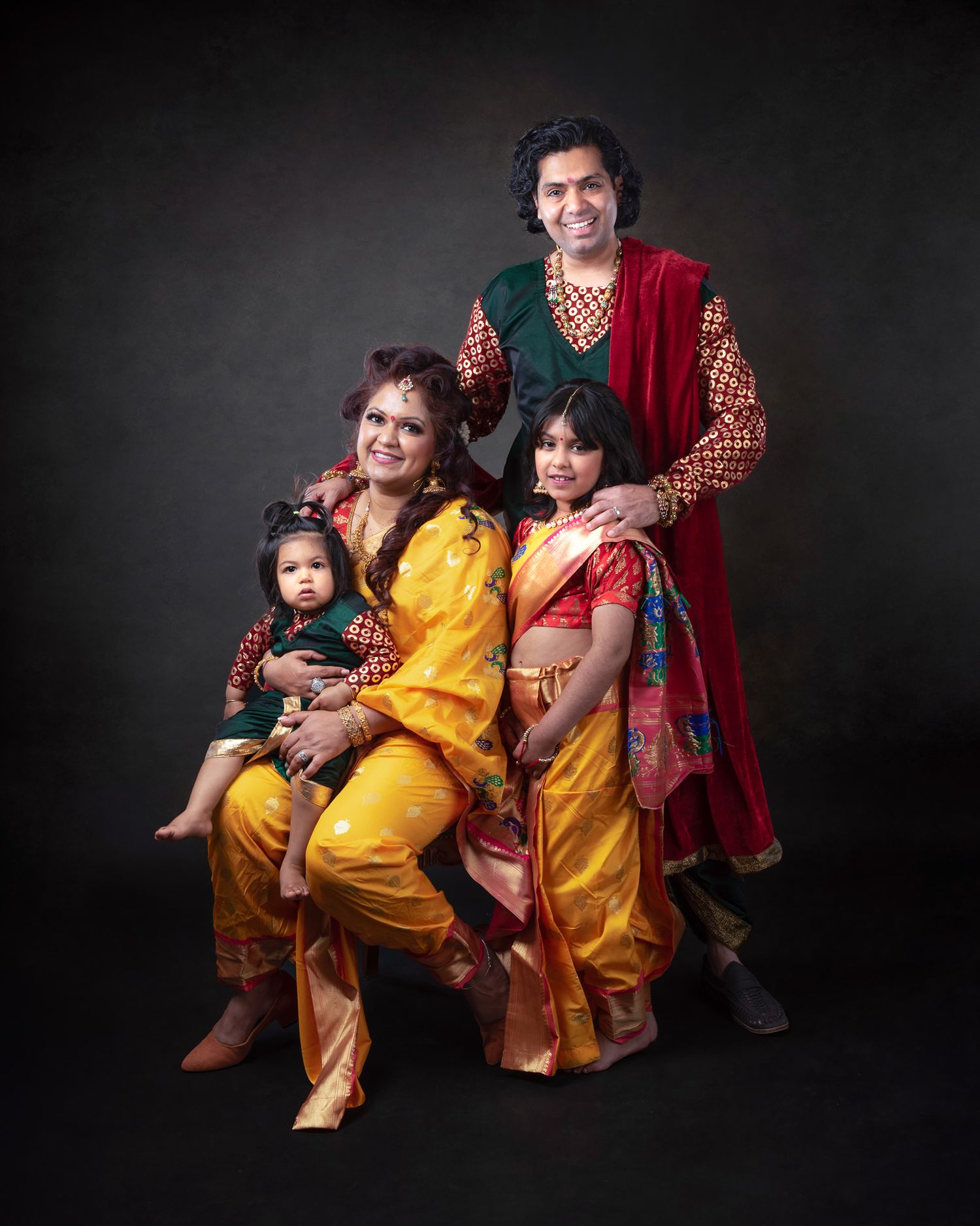 A family poses in traditional Indian clothing against a dark backdrop for a professional portrait session.