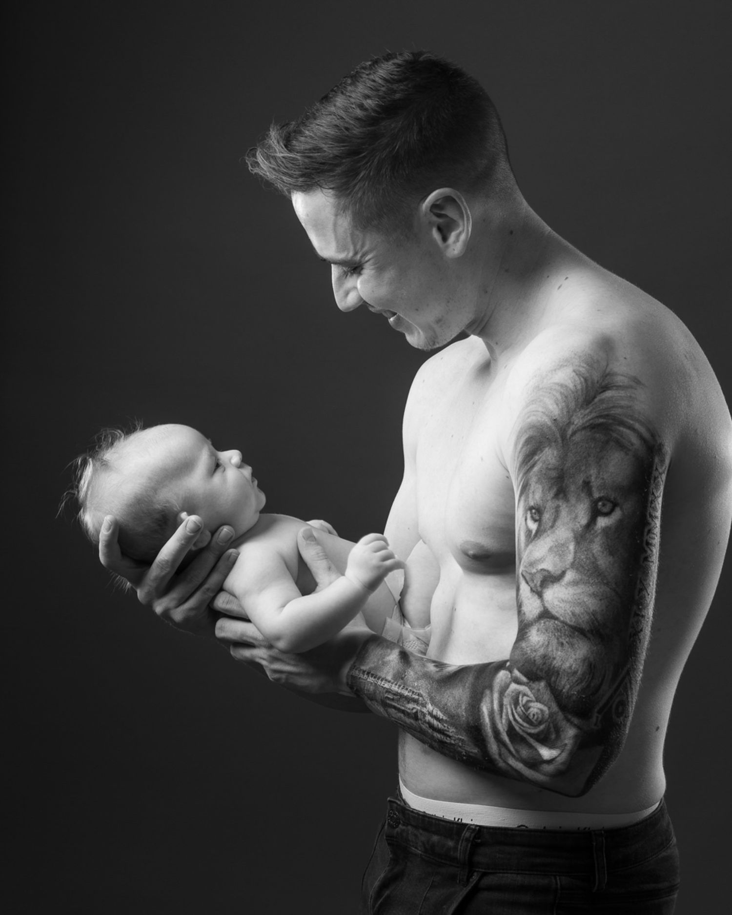 Black and white portrait of a shirtless person with tattoos tenderly holding a newborn baby against dark background.