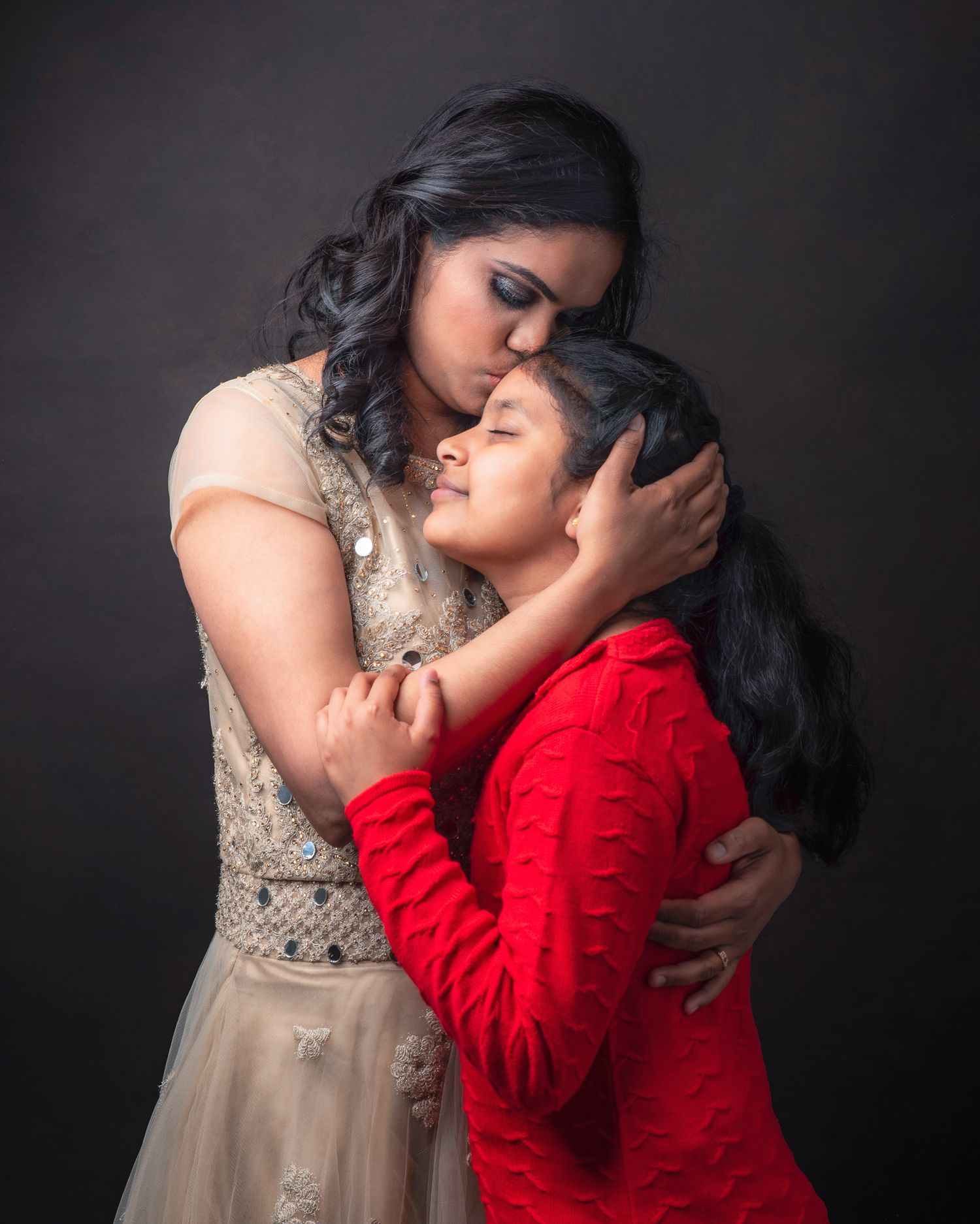 A tender embrace between two figures in elegant attire against a dark backdrop, one in a beige gown and one in red.