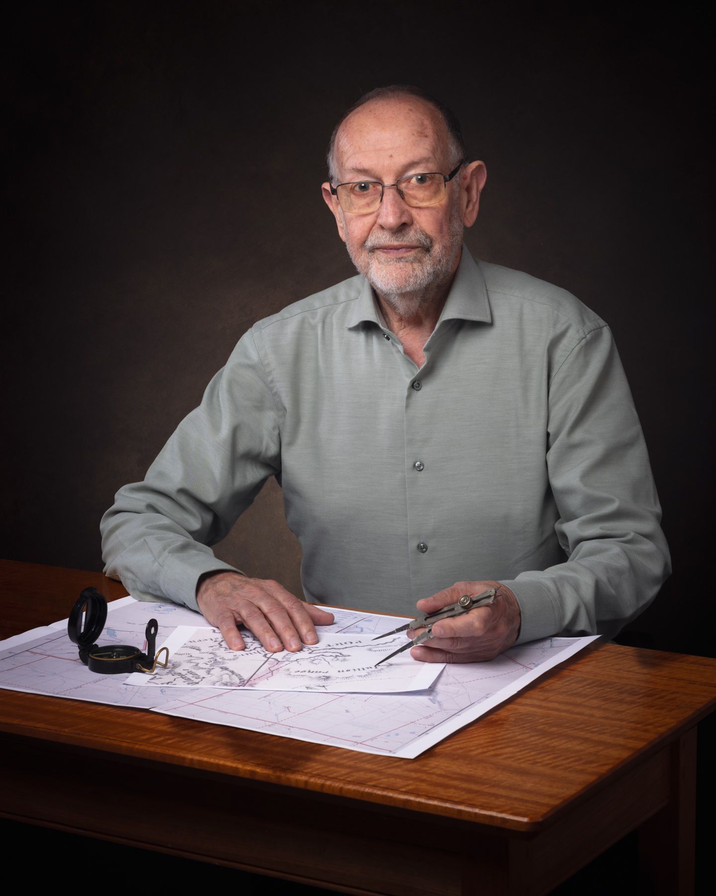 Someone in a gray shirt sits at a wooden desk examining and writing on documents in a dark studio setting.
