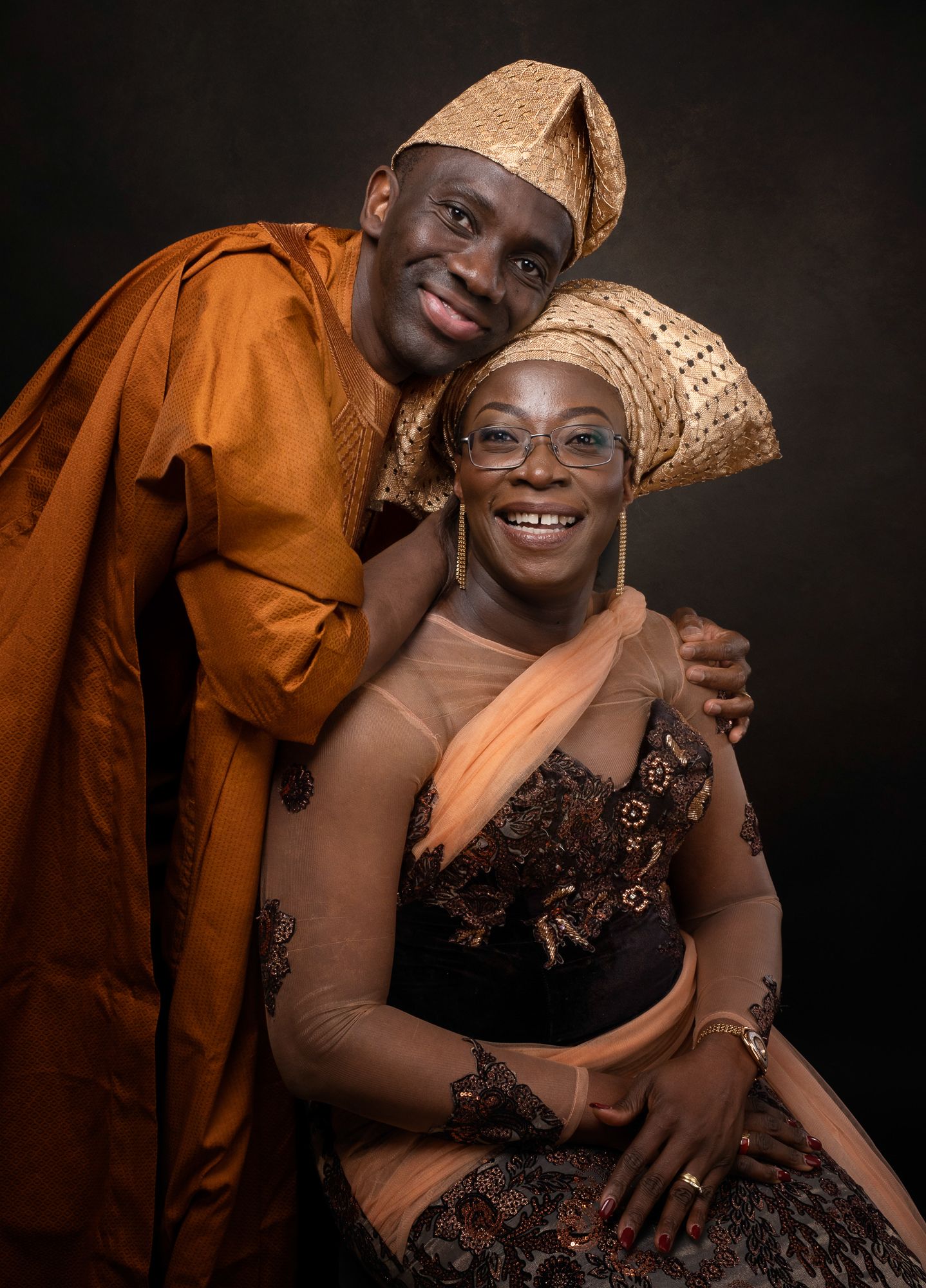 Warm portrait of a couple in traditional African attire with elegant gold headwear against a dark background.