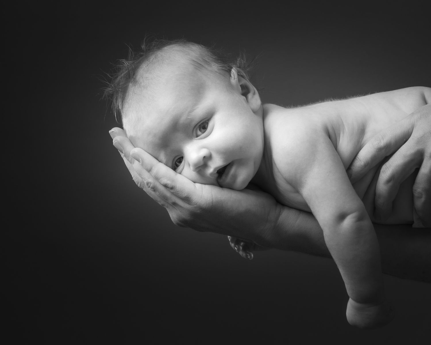 Black and white portrait of a tiny newborn baby cradled gently in adult hands against a dark background.