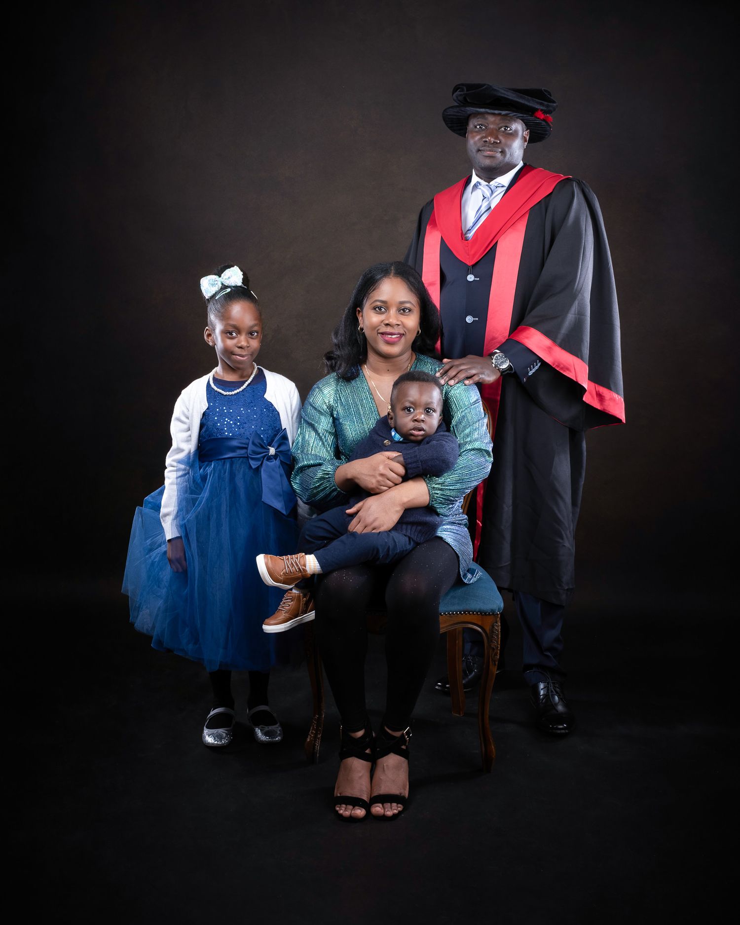 A formal graduation portrait with a graduate in academic regalia seated with family members against a dark backdrop.