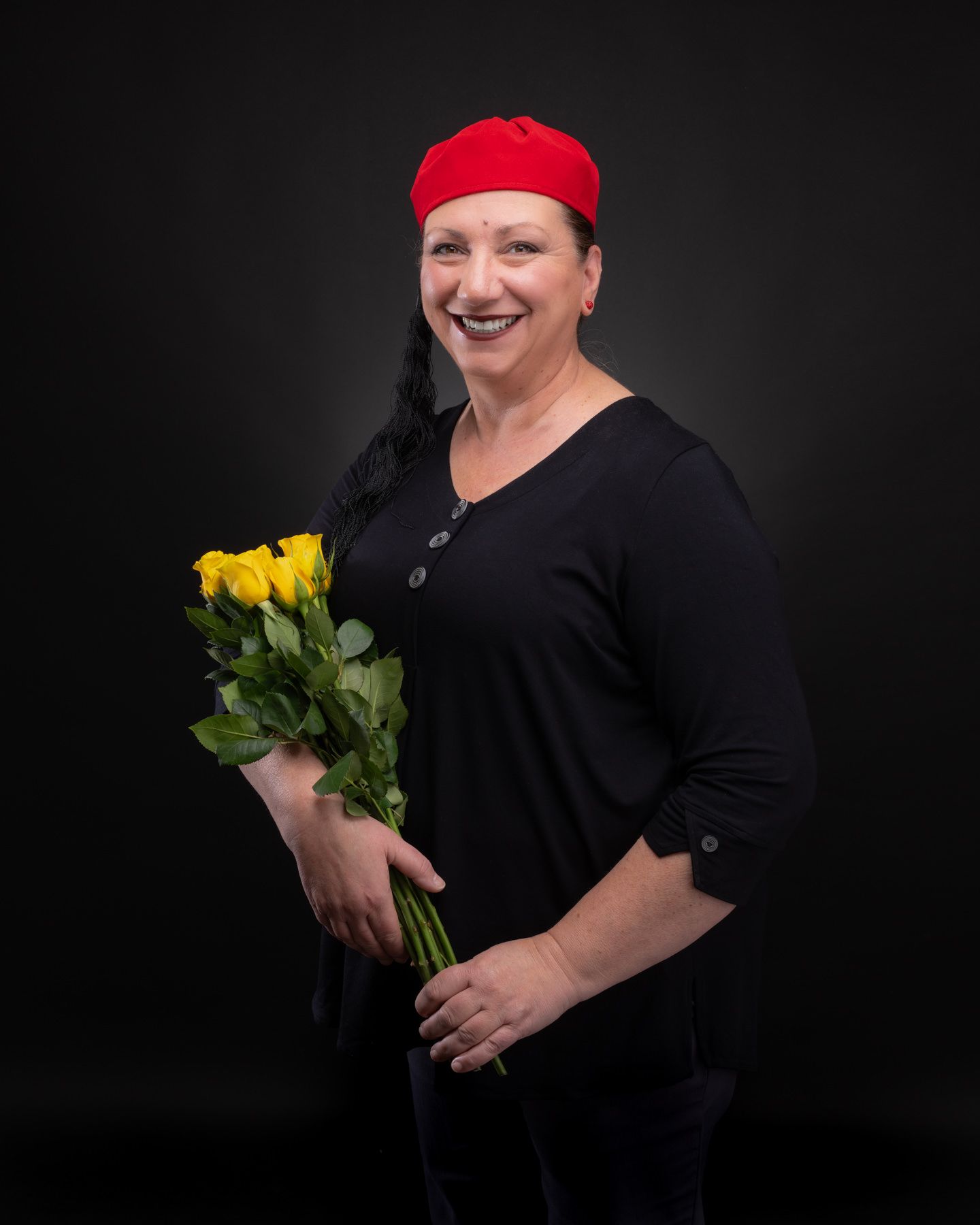 Person in black top and red hat holds vibrant yellow roses against dark background in artistic portrait series.
