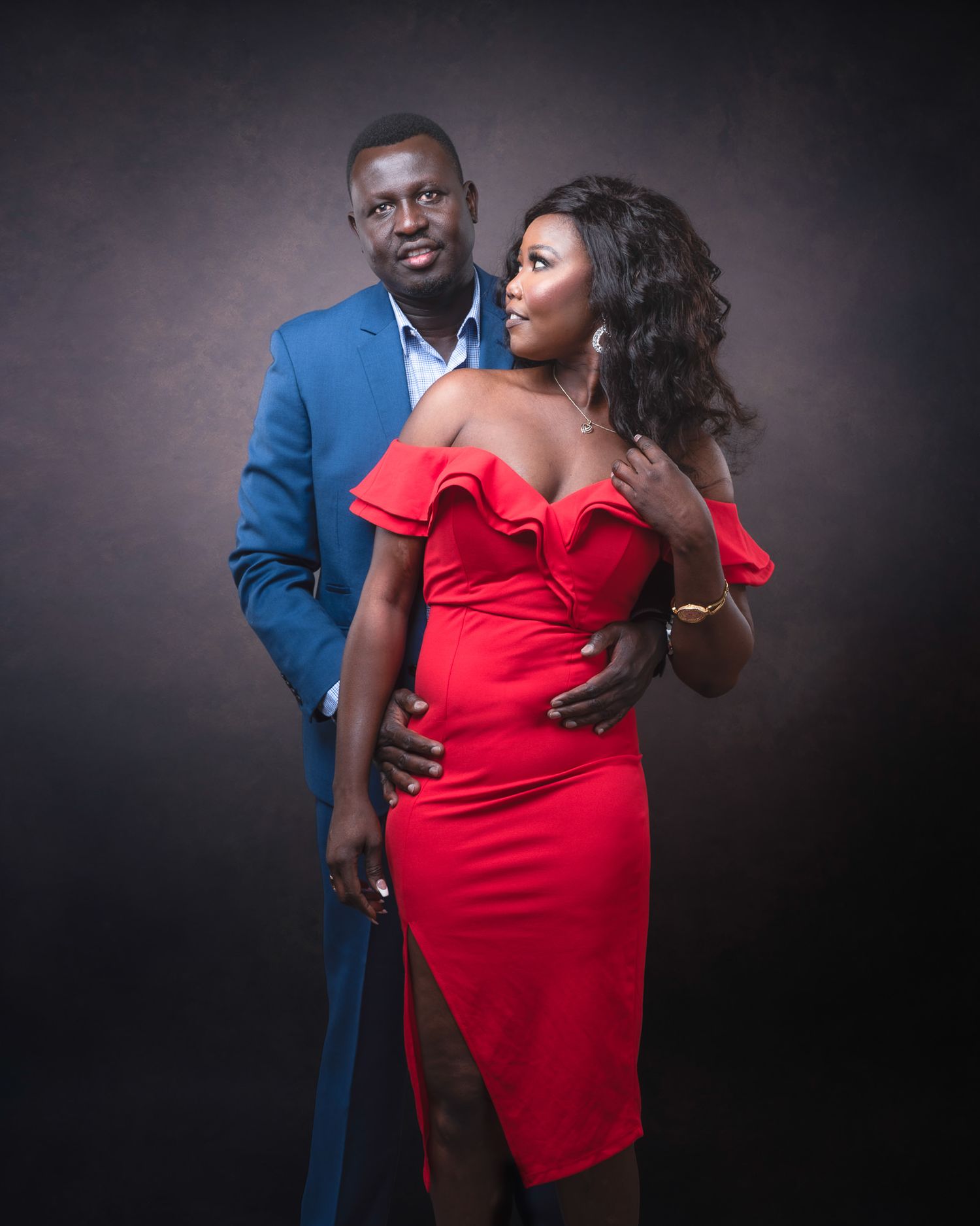 Romantic portrait of a couple posing together, with a blue suit and red off-shoulder dress against a dark backdrop.