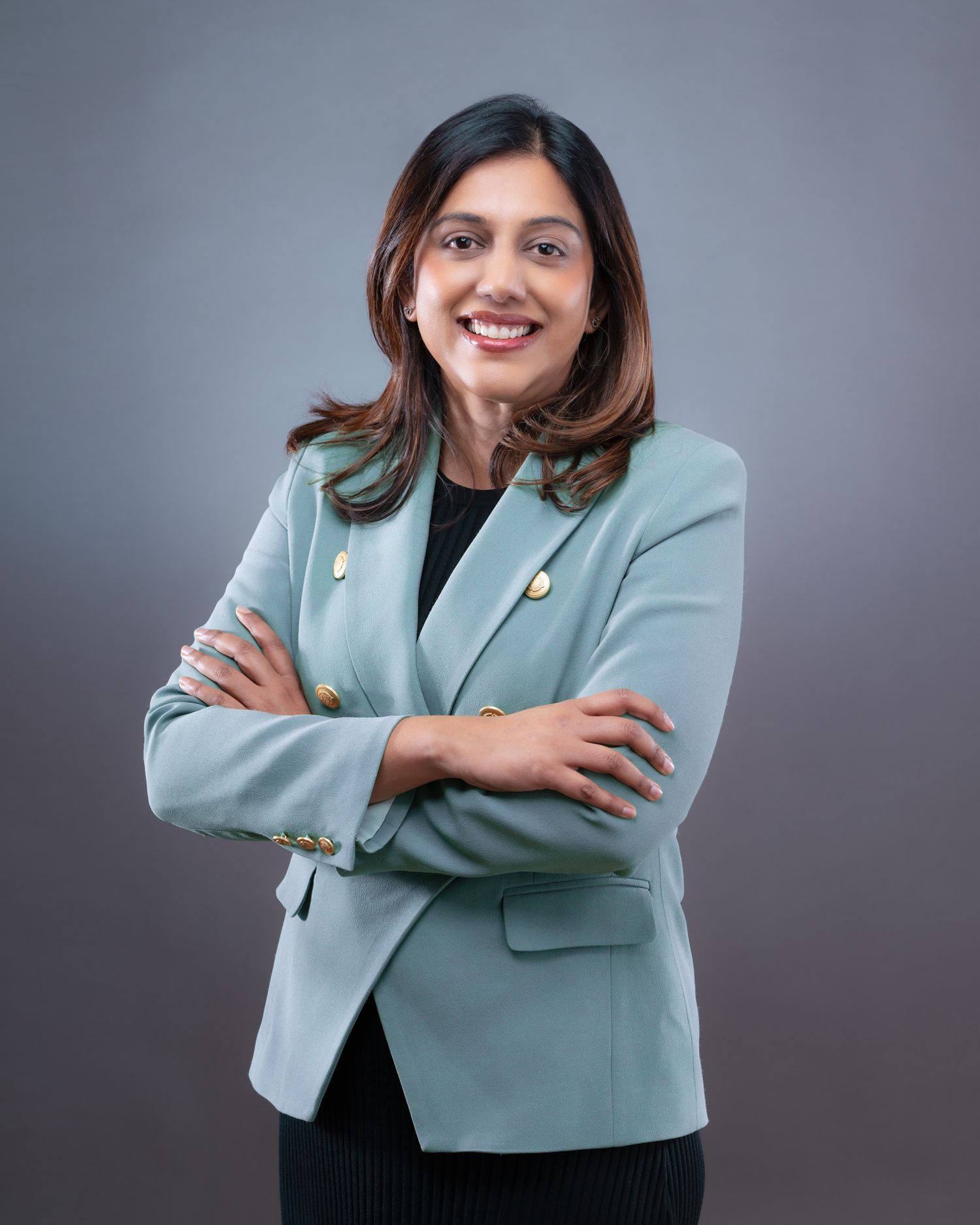Professional wearing a light blue blazer and dark outfit poses against gray background in studio setting.