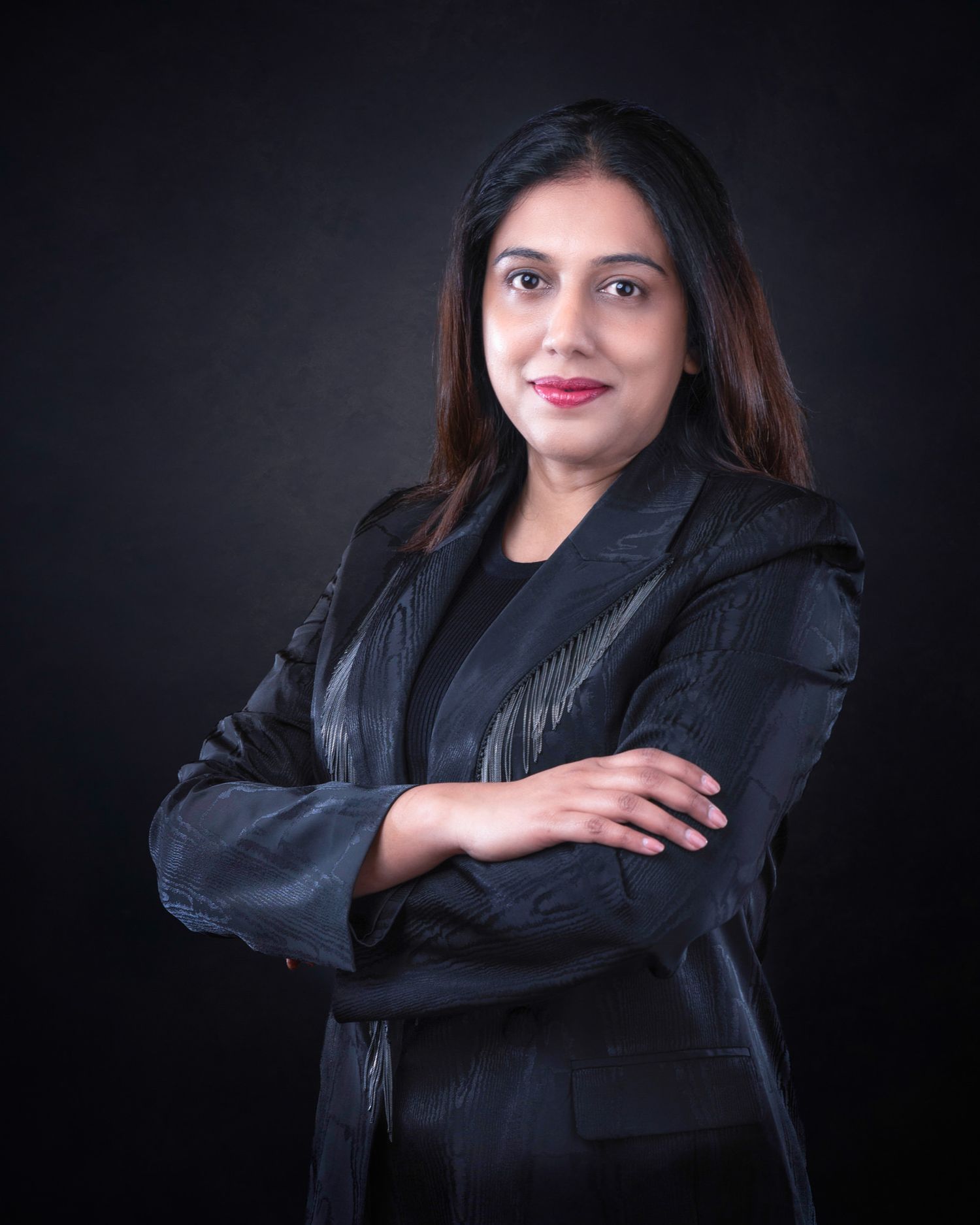 Professional studio portrait of a person in a black blazer and top against a dark background with confident crossed arms.