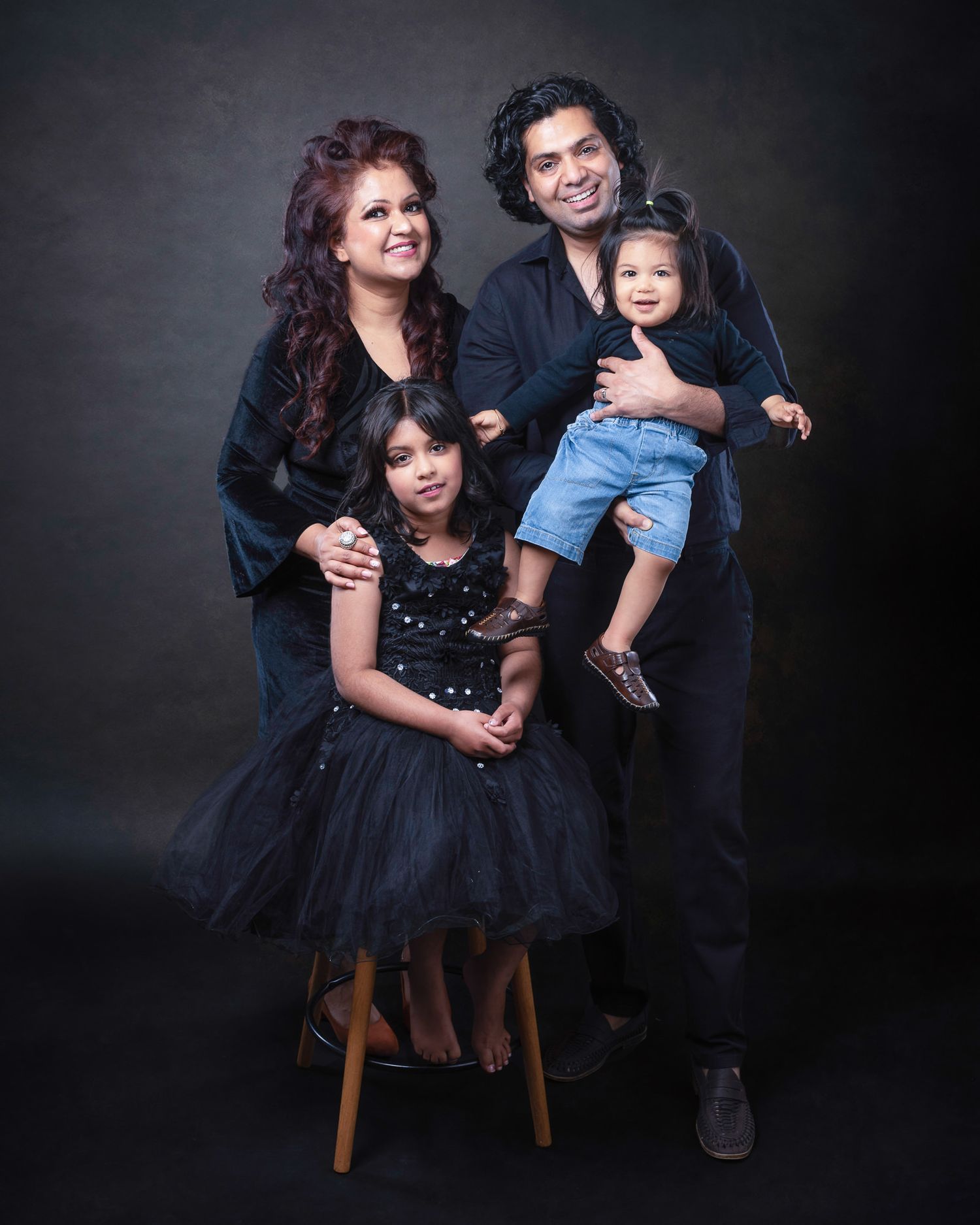 A family portrait in front of a dark backdrop wearing coordinated black outfits in an elegant studio setting.
