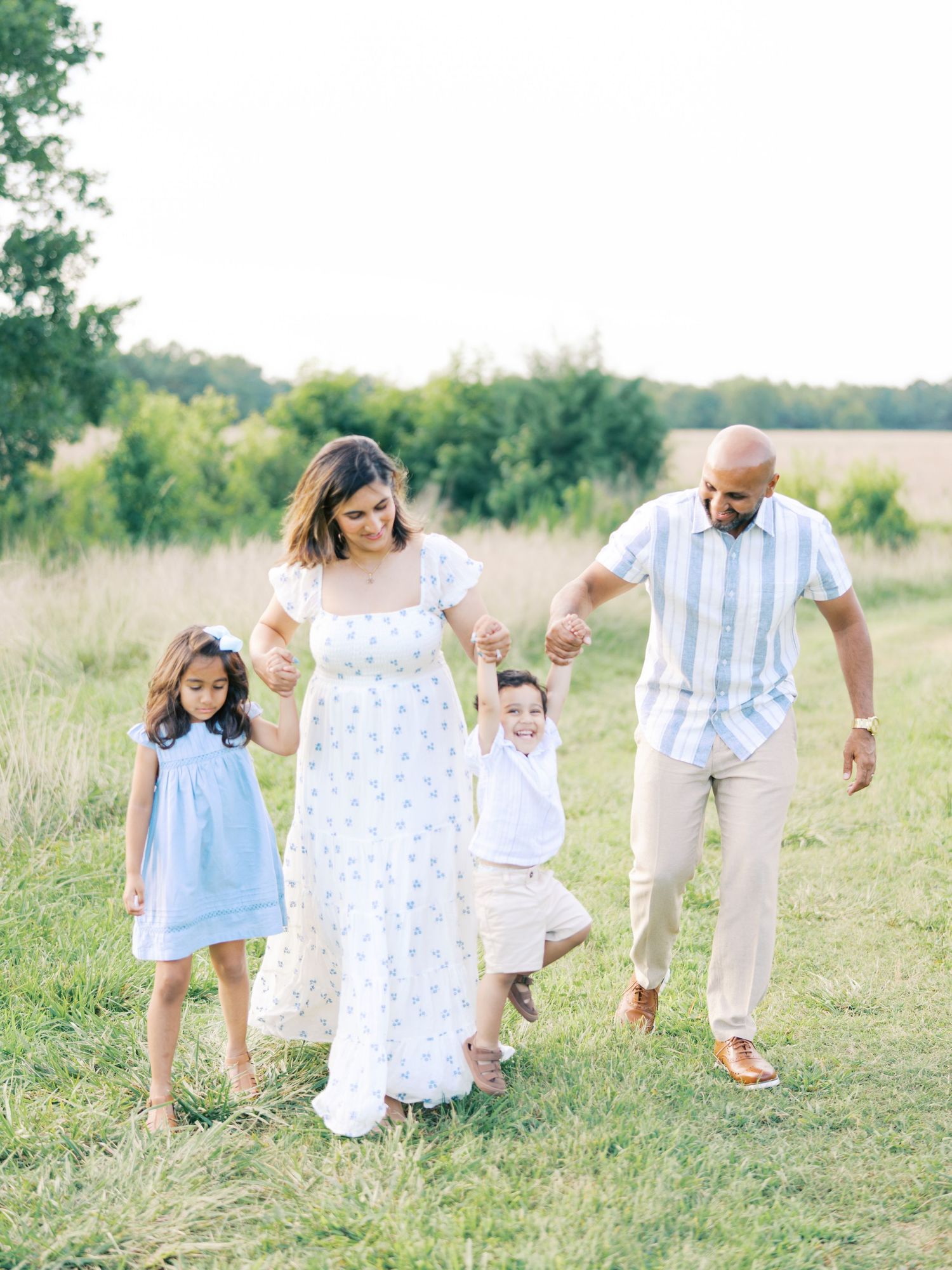 Family in coordinating white and blue outfits walks together through a sunlit meadow on a summer evening.