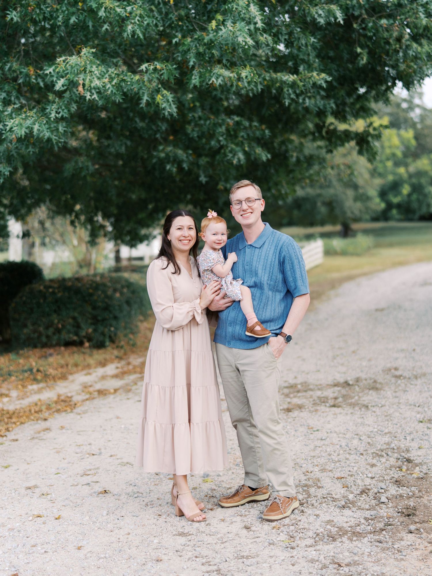 A family in coordinating pink and blue outfits poses together on a gravel path surrounded by green trees.