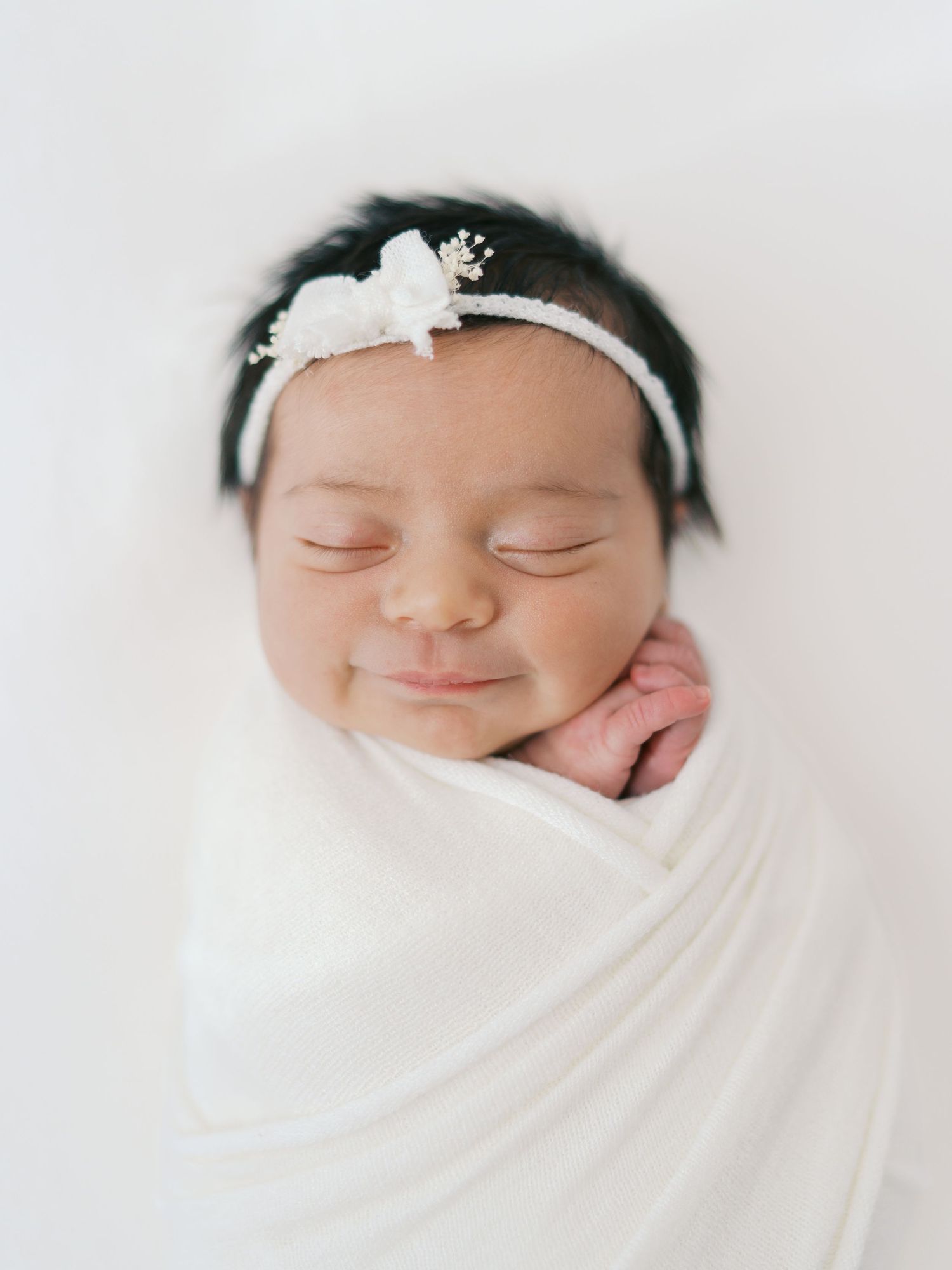 Smiling newborn baby girl swaddled in white wrap wearing a delicate white bow headband, sleeping peacefully.
