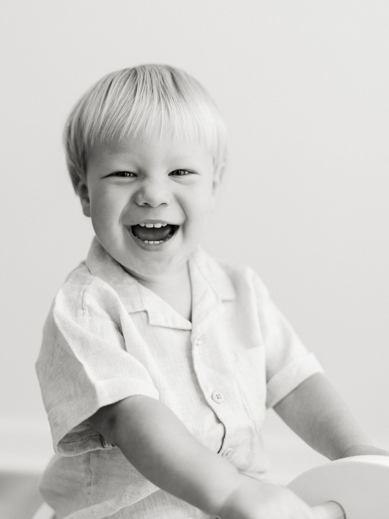 Black and white portrait of a laughing blonde toddler boy in a linen shirt, radiating joy.