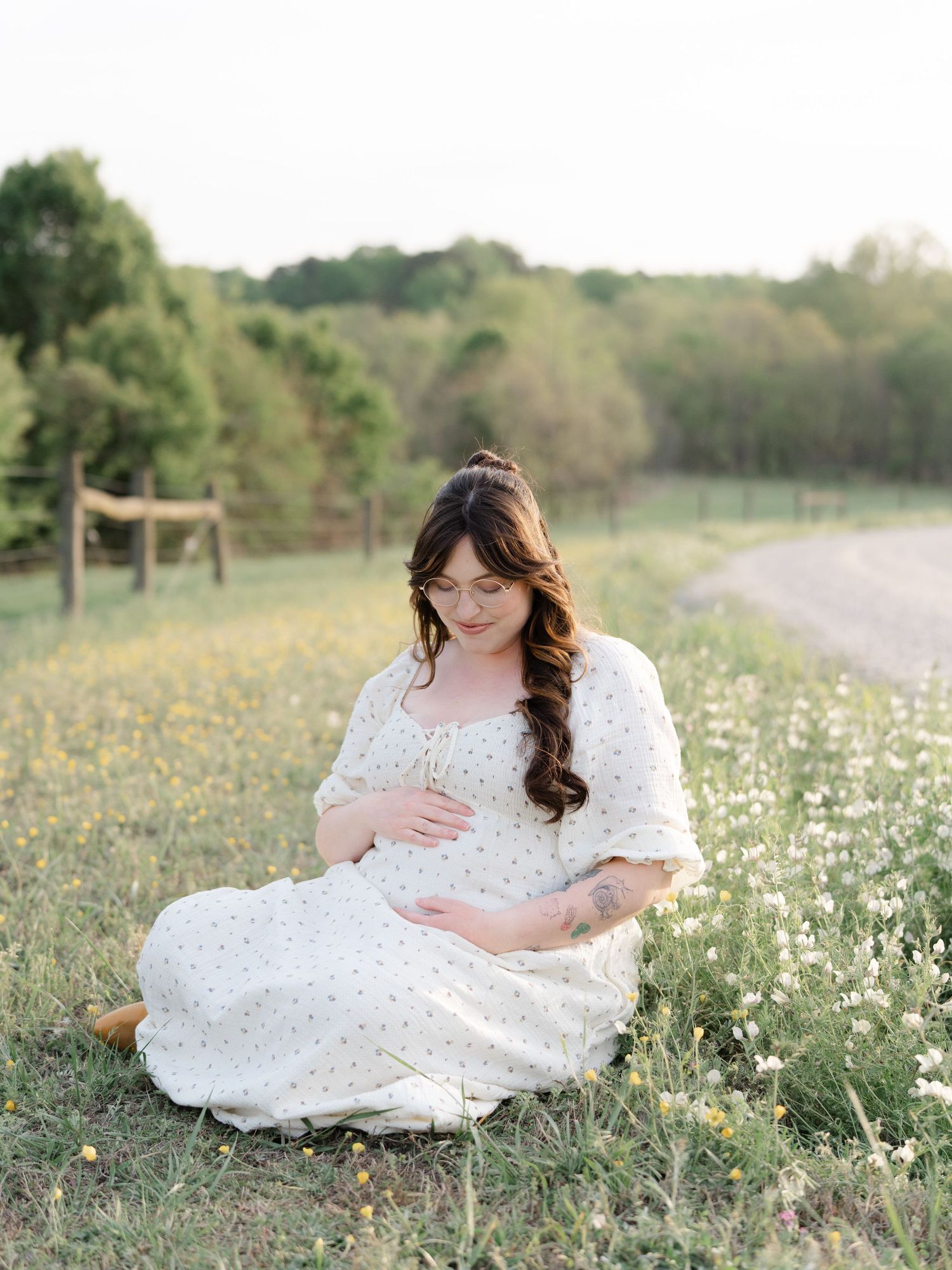 Pregnant woman in white floral dress sitting in wildflower field during golden hour maternity photoshoot.