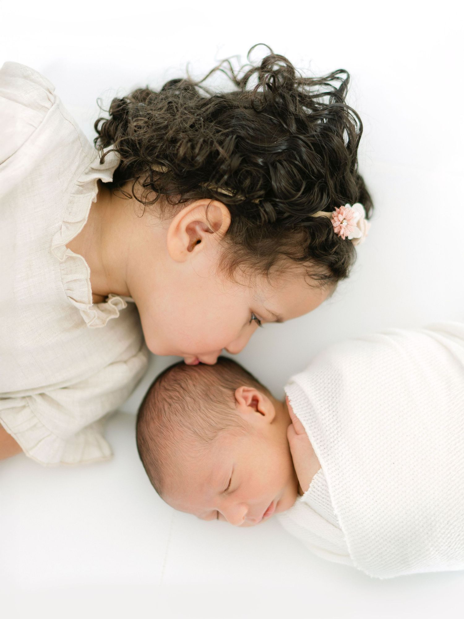 A young girl with curly hair gently kisses a sleeping swaddled newborn baby on a white background.