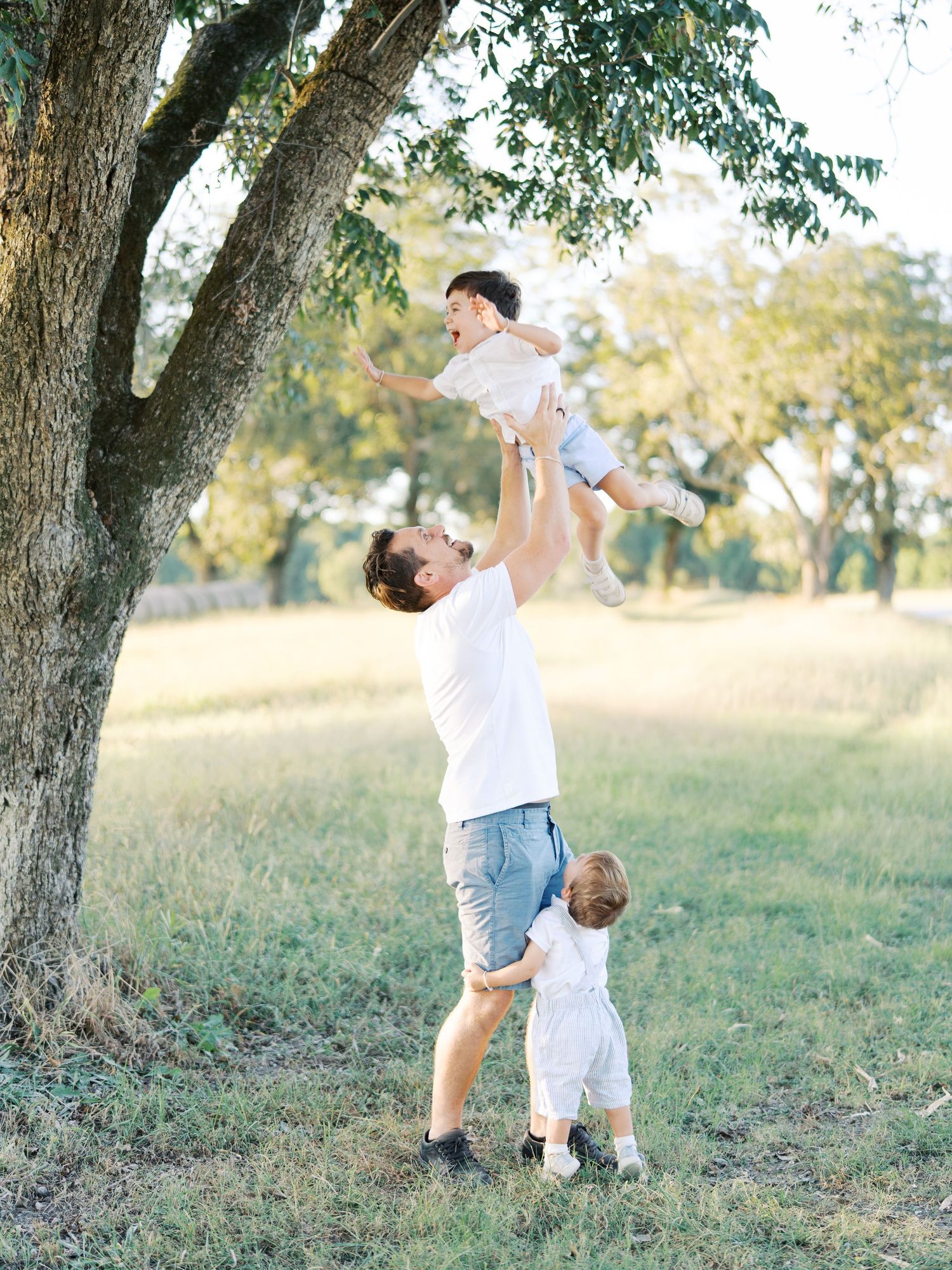 Playful parent and child in white outfits dance together under a large tree in a sunny outdoor field.