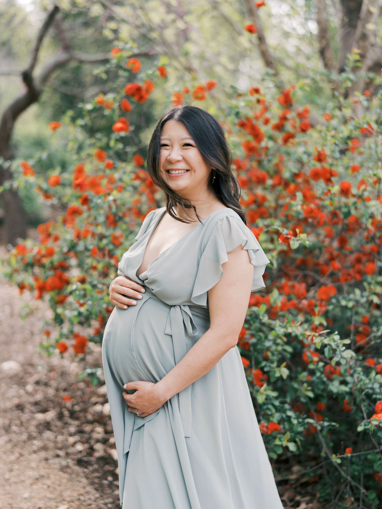 Smiling expectant mother in grey dress stands against backdrop of bright orange flowers.