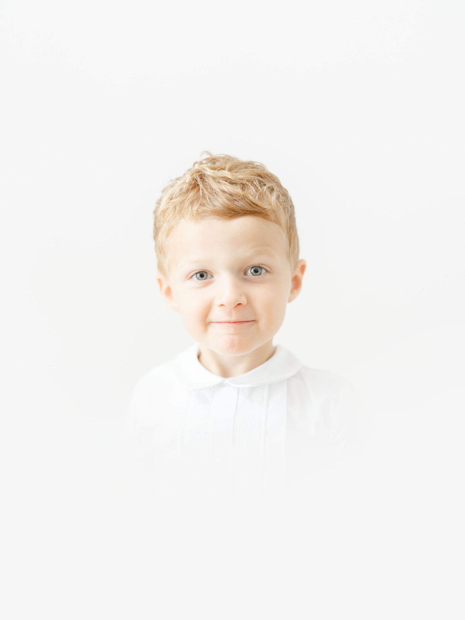 Smiling young blonde boy with blue eyes wearing a white shirt against a bright white background.
