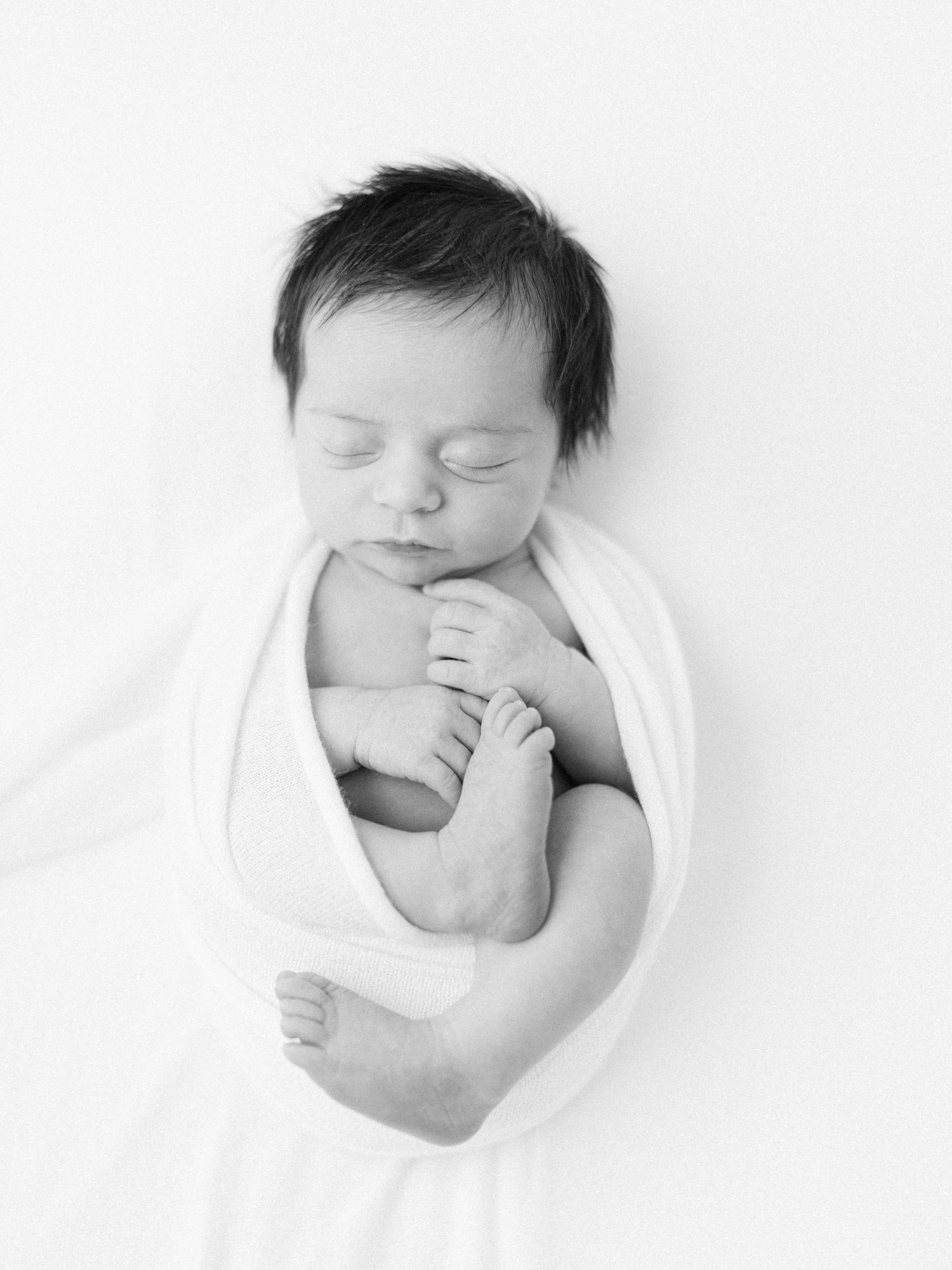 Black and white newborn photo of a sleeping baby swaddled in white wrap, peacefully resting with tiny hands and feet visible.