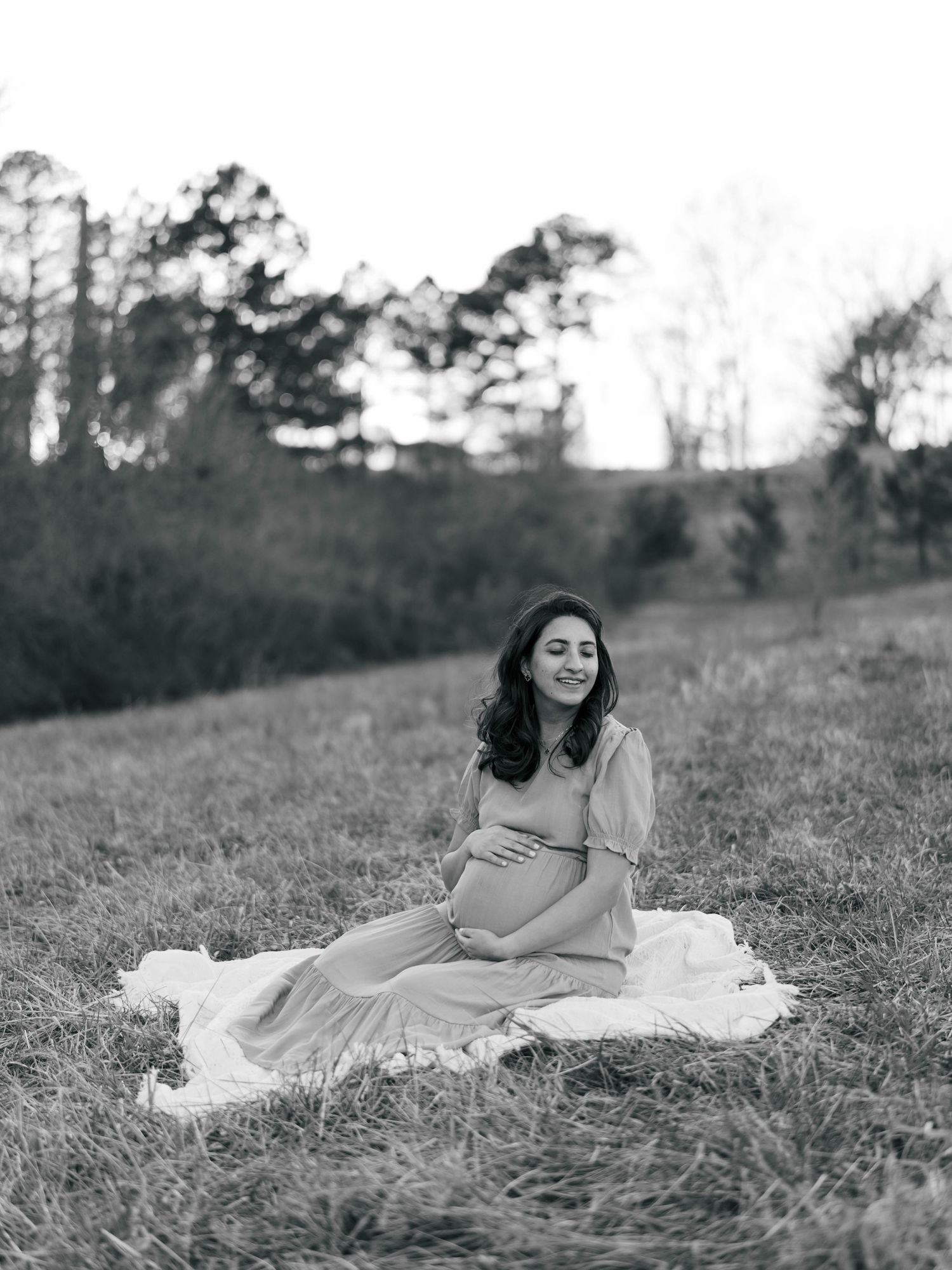 Black and white maternity photo of a woman in a flowing dress sitting on a blanket in a grassy field.
