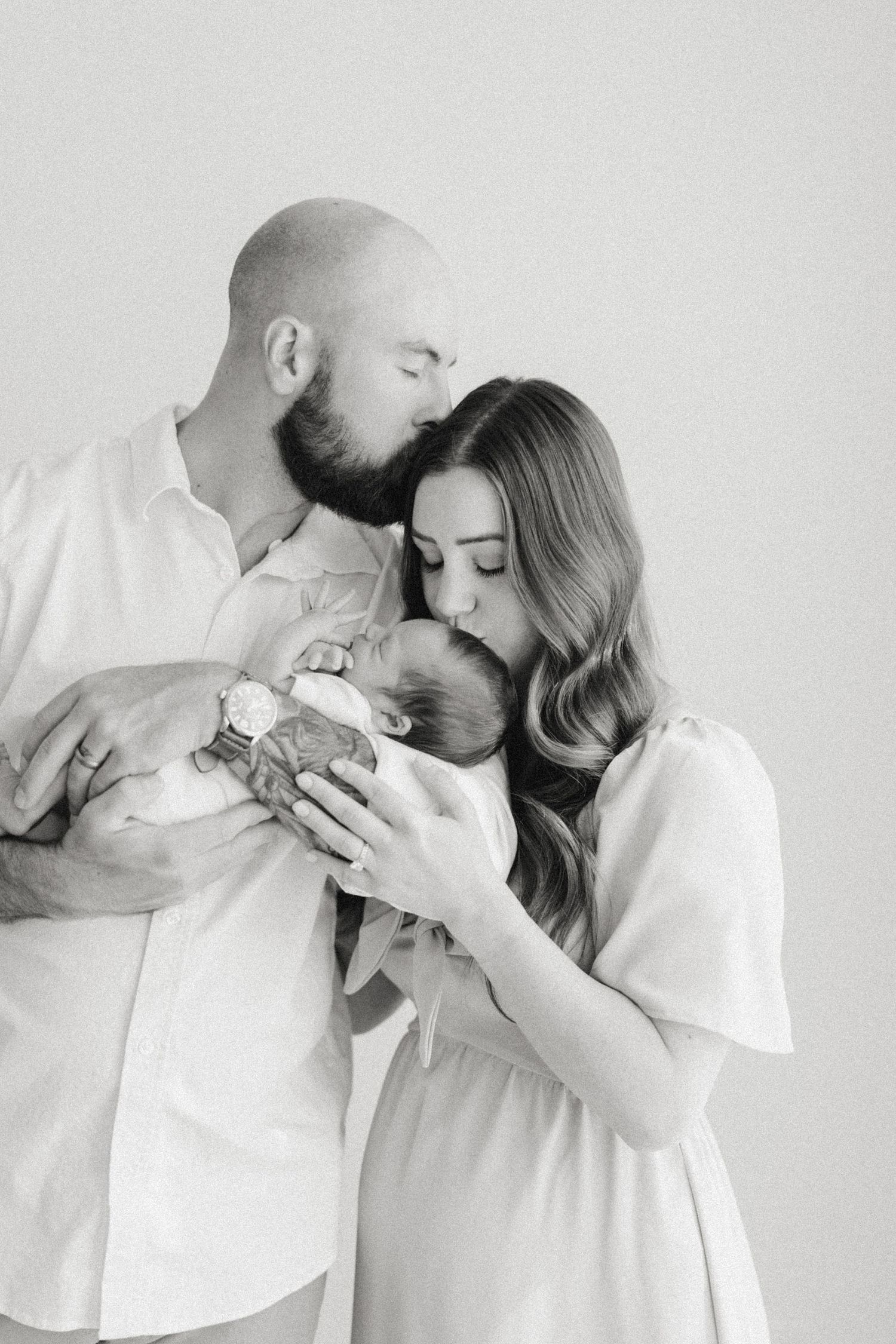 Black and white photo of parents kissing their newborn baby in a tender studio portrait after visiting baby boutiques in Raleigh.