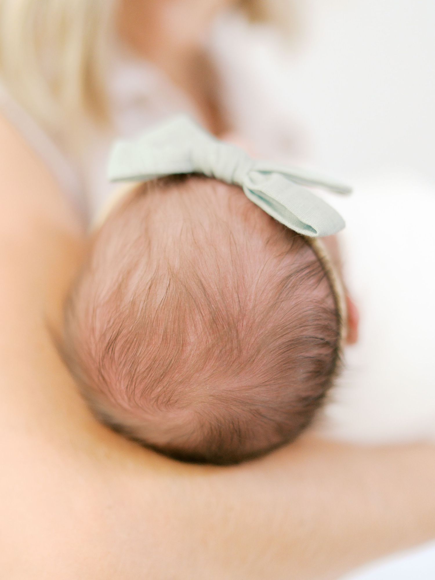 Close-up of a newborn baby's head with a sage green bow headband, cradled in mother's arms.