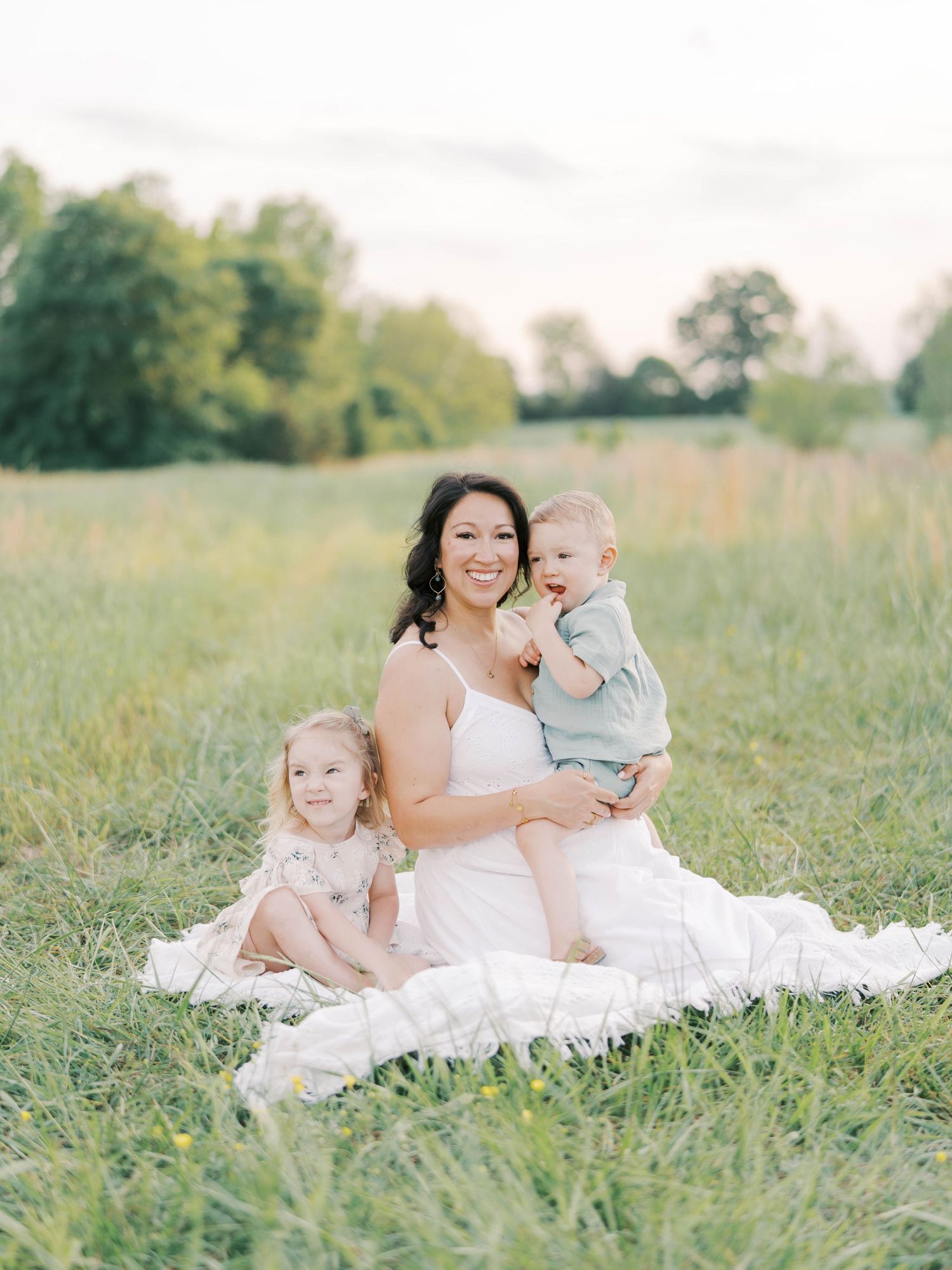 Maternity portrait with children sitting on white blanket in green field.