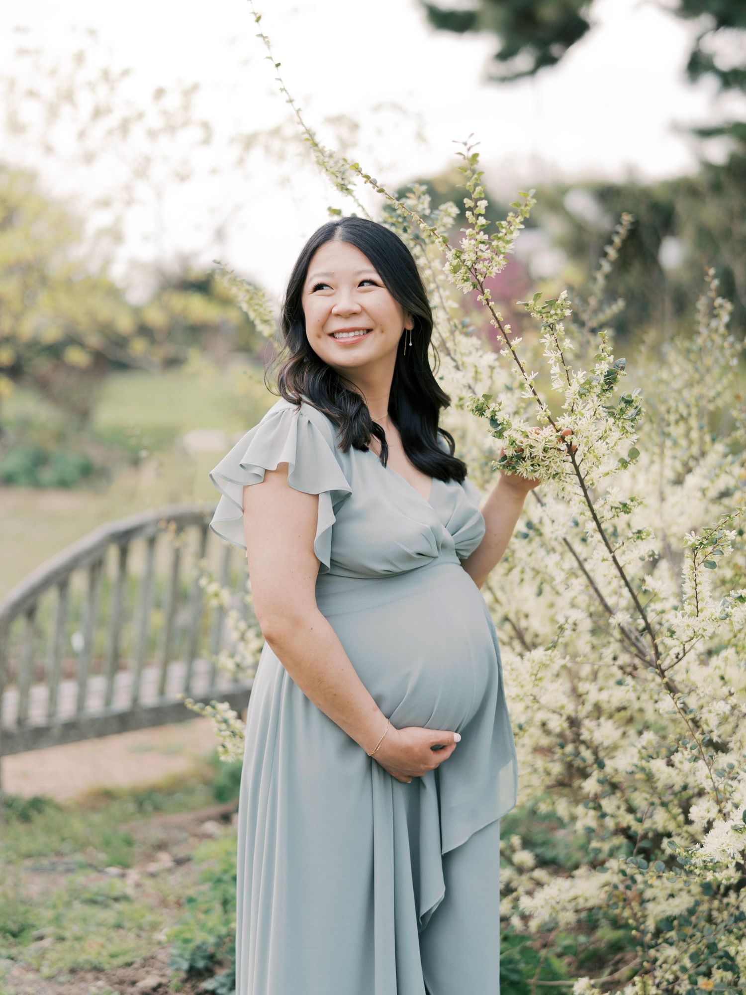 An expectant mother in a flowing blue-gray dress poses near blooming cherry blossoms in a garden setting.
