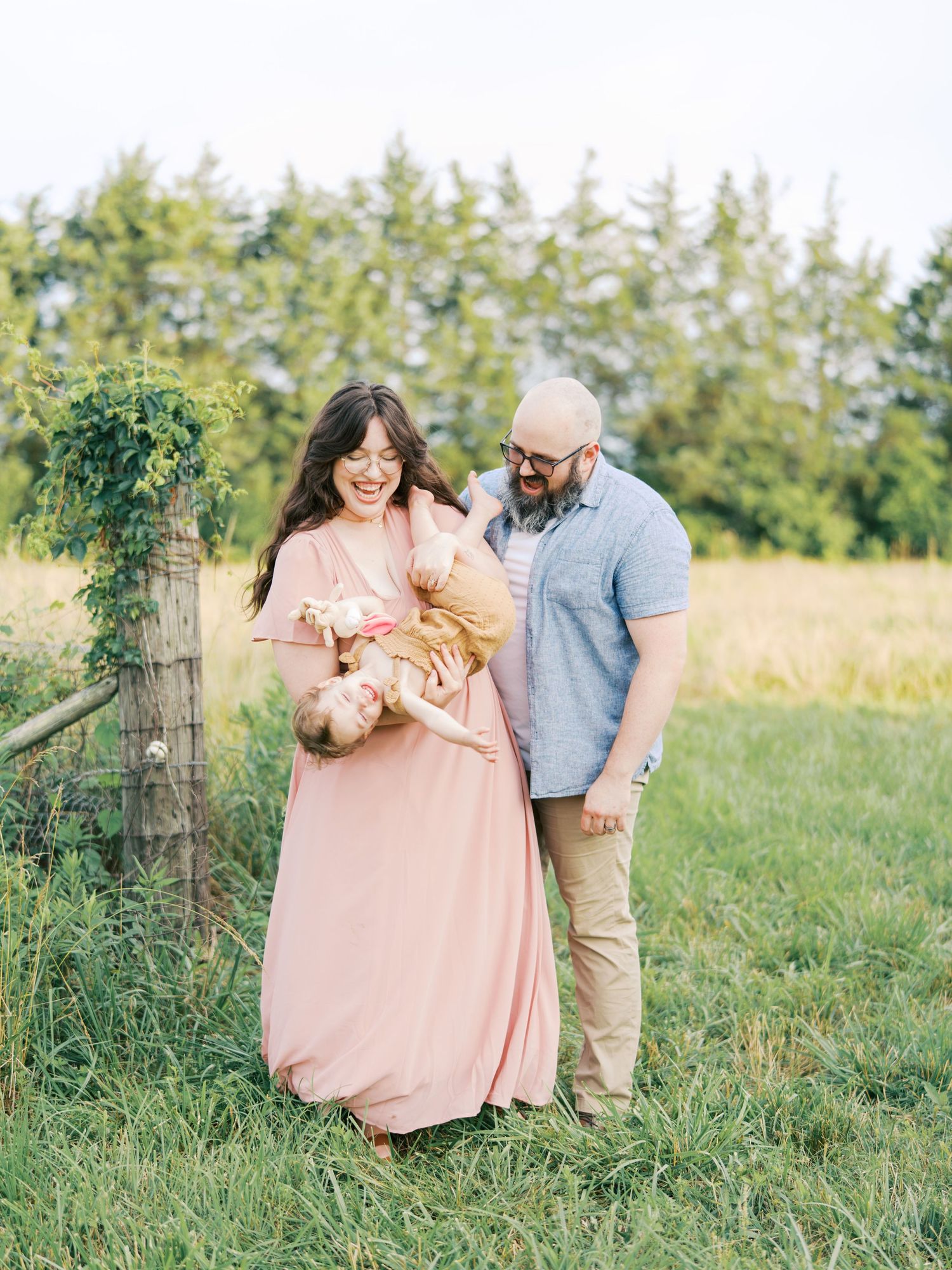 A joyful family of three poses in a green field, mother in pink dress holding baby, father in blue shirt smiling nearby.
