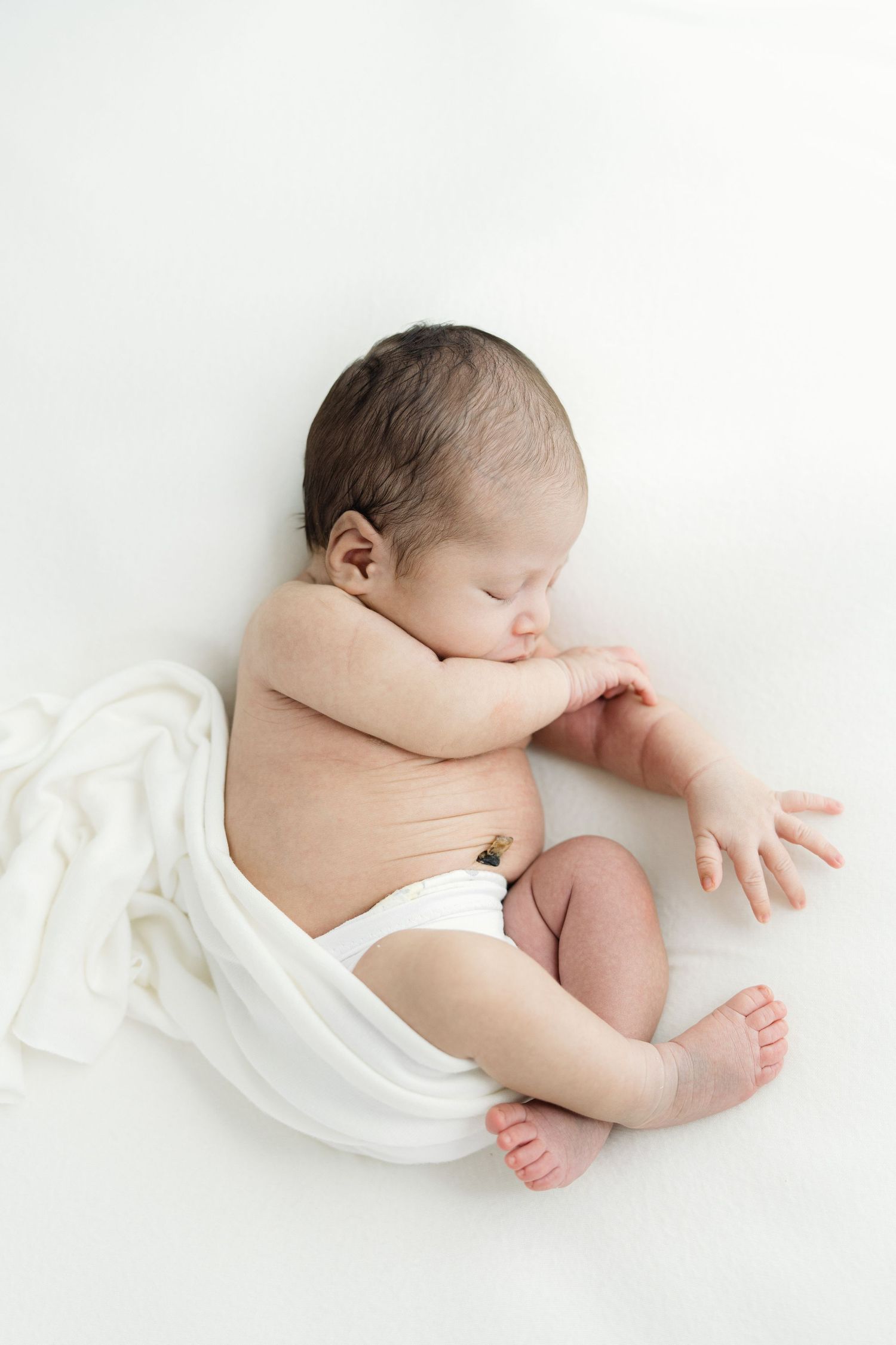 Newborn baby sleeping peacefully on white background, wrapped in white cloth with dark hair and tiny hands visible.