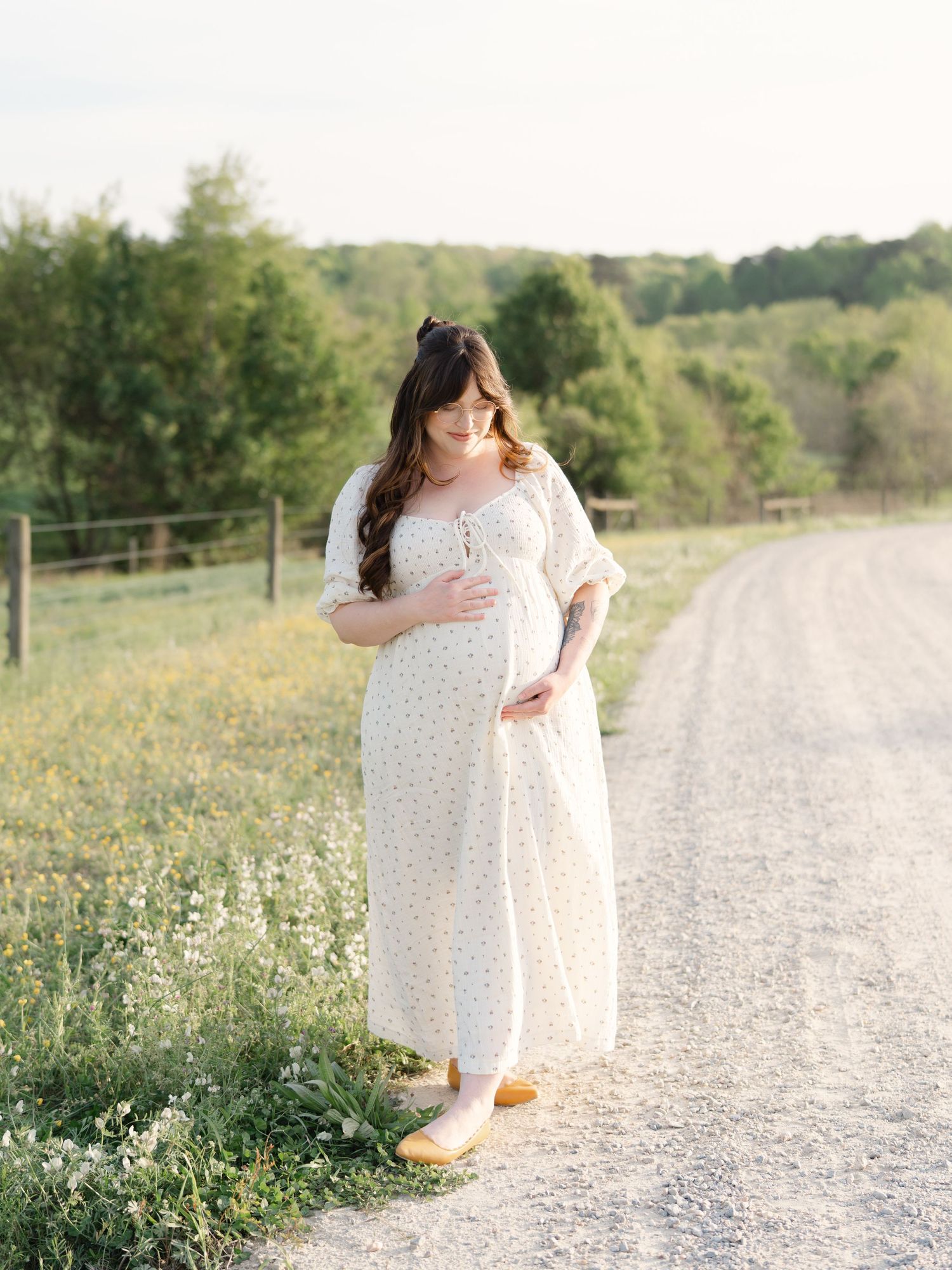 Pregnant woman in floral maxi dress standing on rural gravel road surrounded by green fields at golden hour.