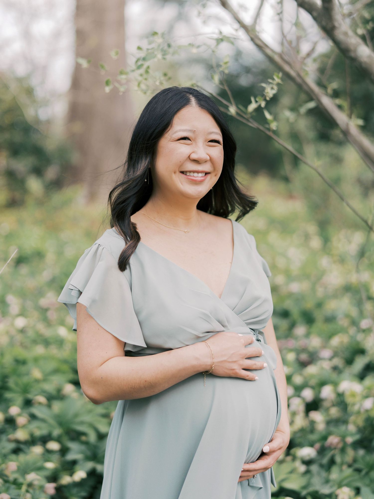 An expectant mother in a flowing gray dress smiles radiantly in a spring garden setting.
