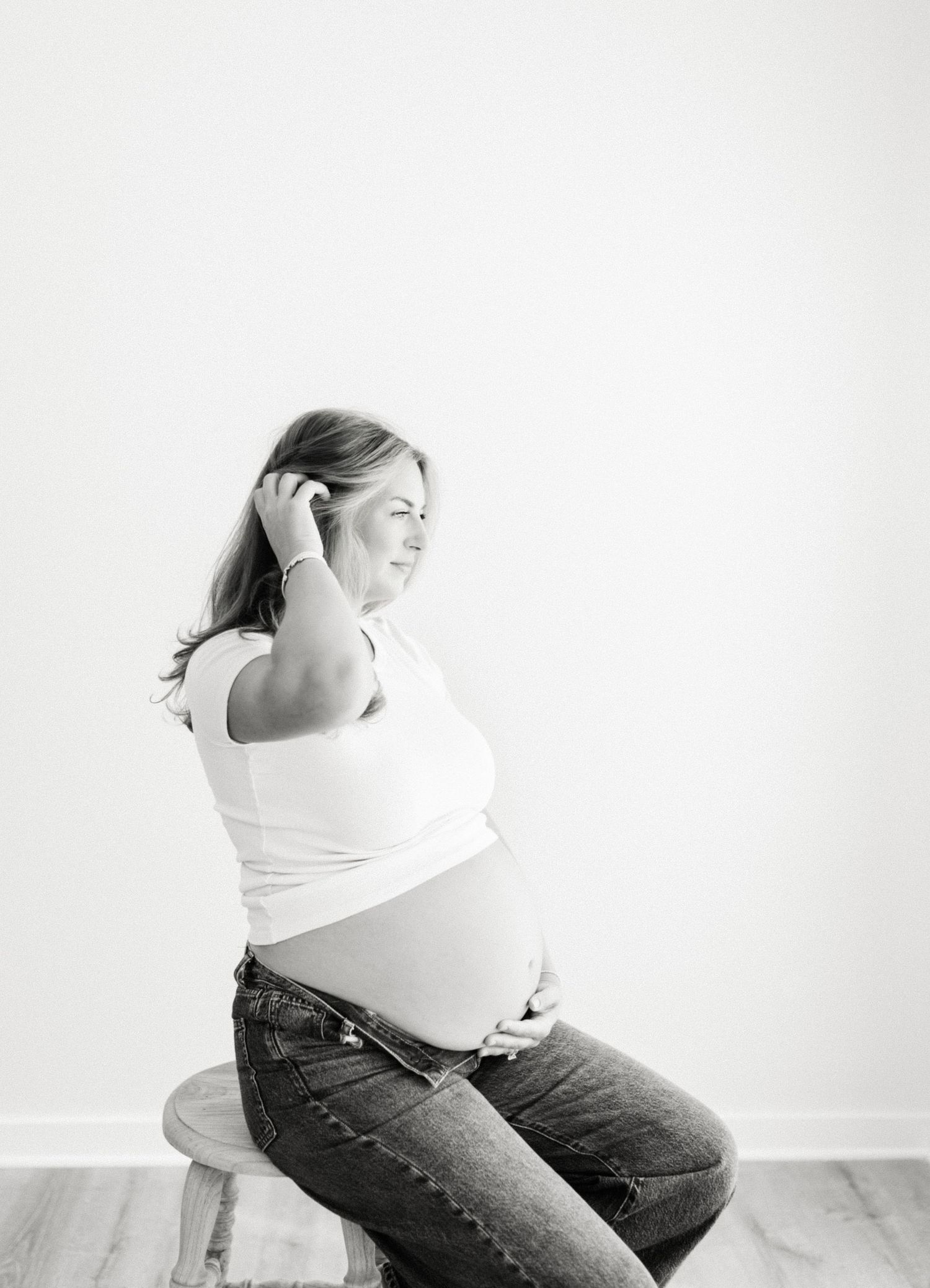 Black and white photo of a pregnant woman sitting, cradling her bare belly while tucking hair behind her ear.