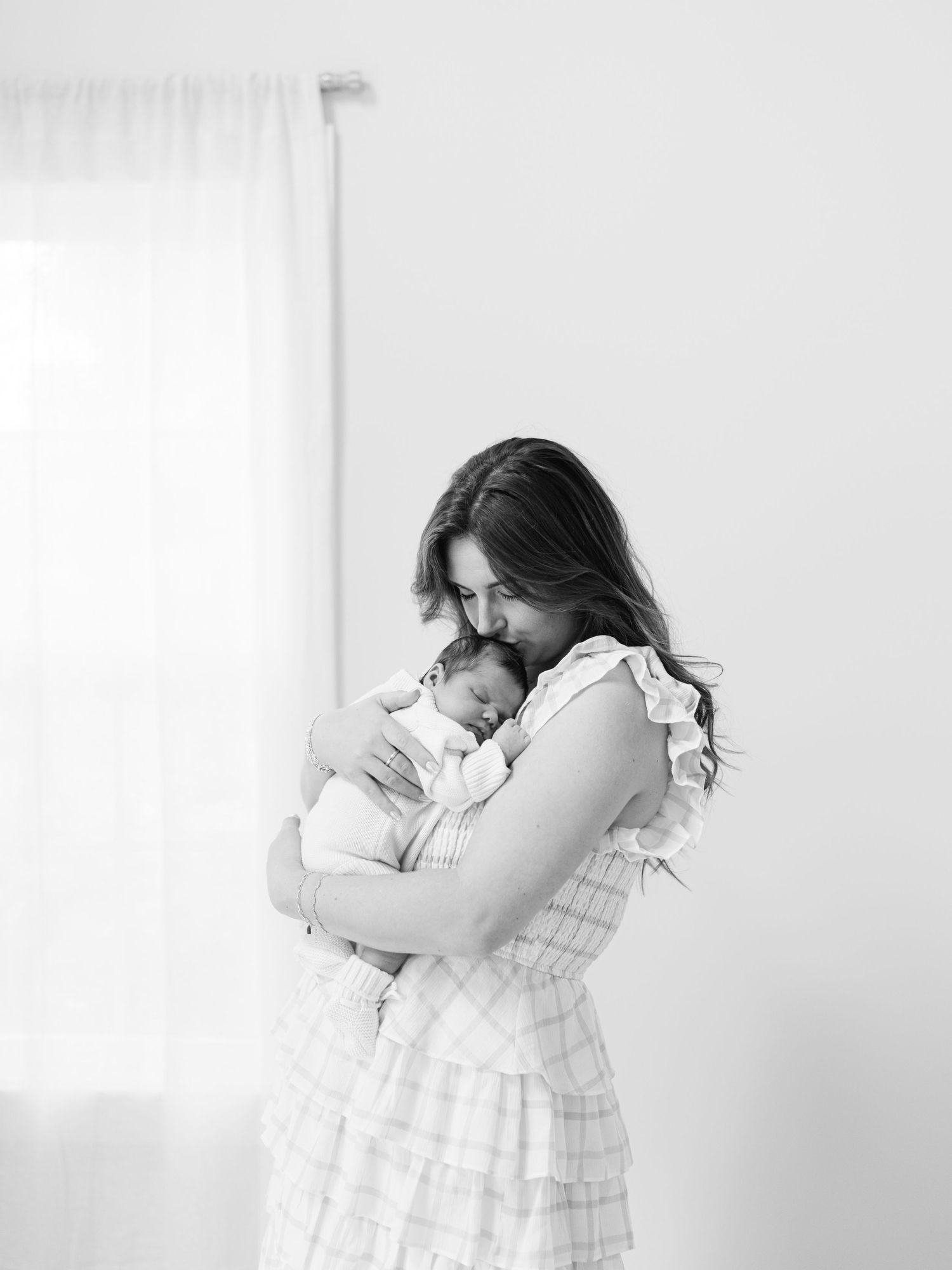 Black and white photo of a mother tenderly cradling her newborn baby near a bright window.