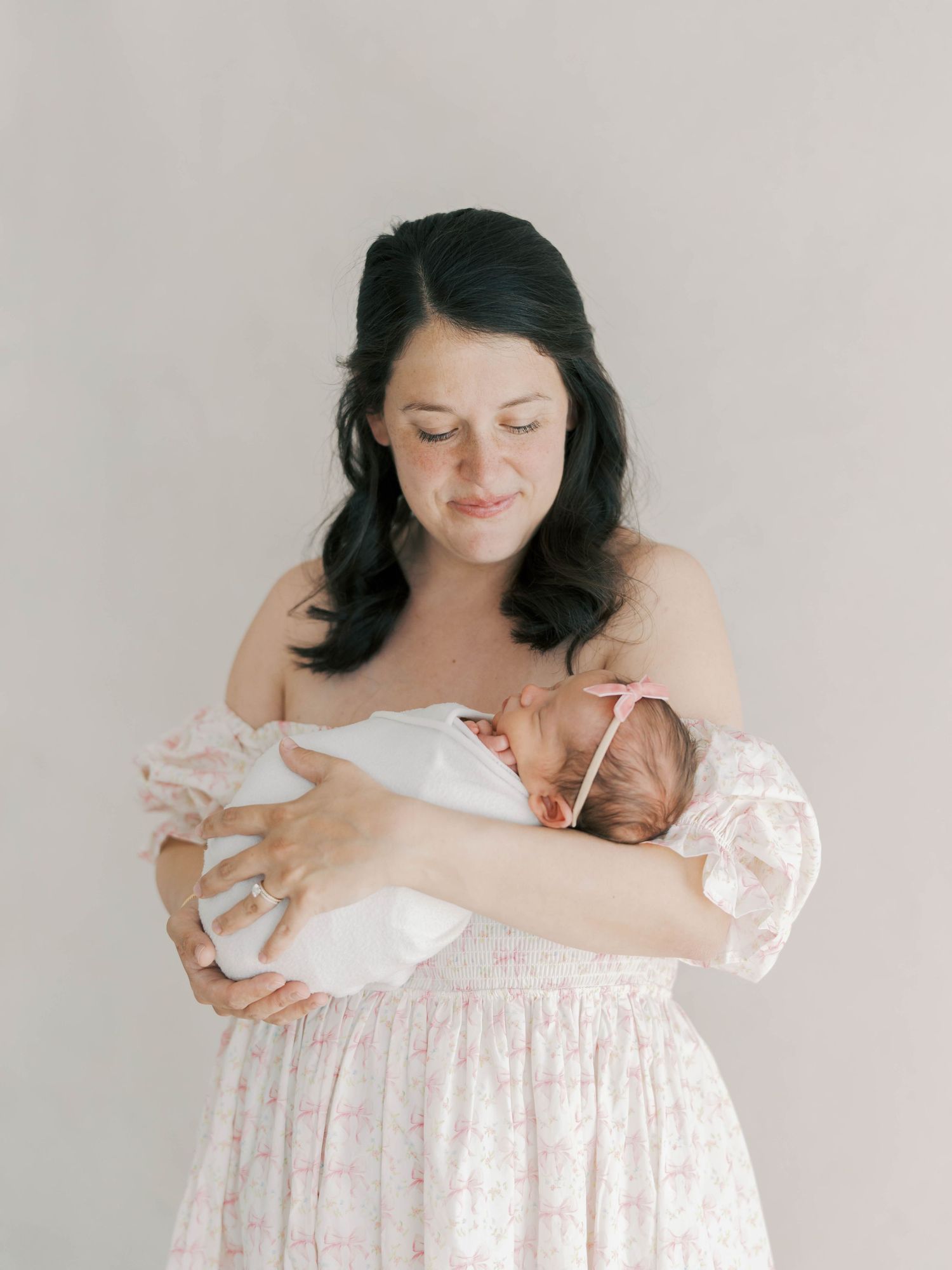 A dark-haired mother in a floral pink dress tenderly holds her swaddled newborn baby in a soft studio portrait.