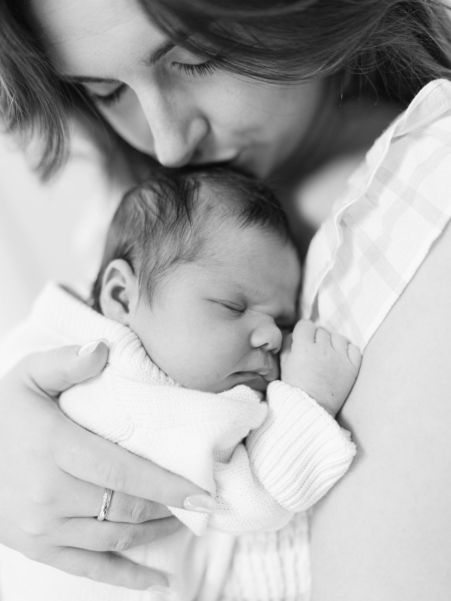 Mother gently kisses her sleeping newborn baby in a tender black and white portrait photo.